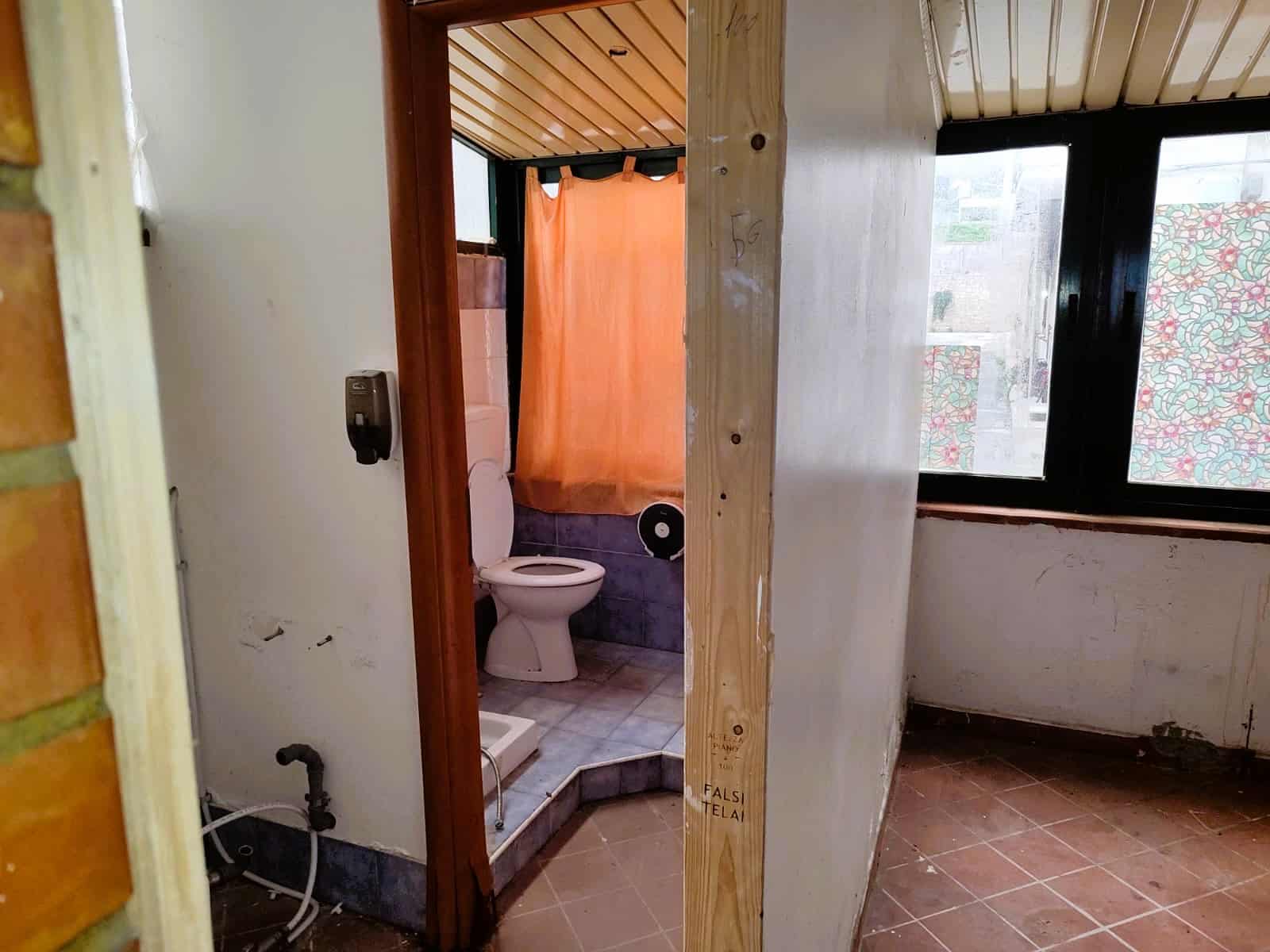 Bagno da ristrutturare in appartamento, con pareti in intonaco grezzo e pavimento in piastrelle, finestra grande con tende colorate, areas di lavoro per ristrutturazione.