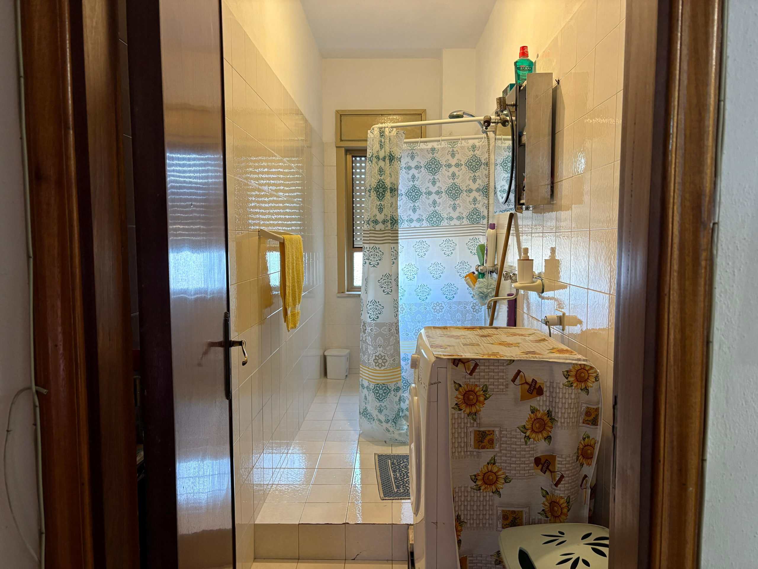 Lavanderia con lavabo, lavatrice, servizi igienici, tenda da doccia decorativa e finestra, spazio funzionale e ben illuminato, ideale per la gestione domestica.