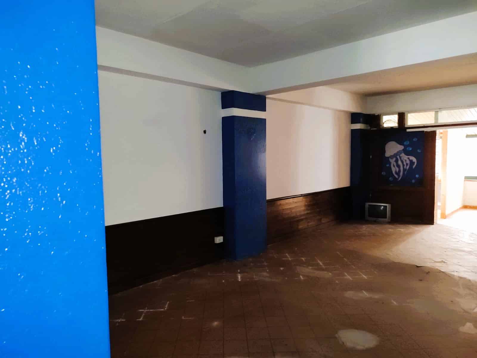 Immagine di un soggiorno vuoto con pareti bianche, una parete decorata con un quadro di un polpo e un televisore, pavimento in piastrelle marroni, e grande finestra che lascia entrare molta luce naturale.