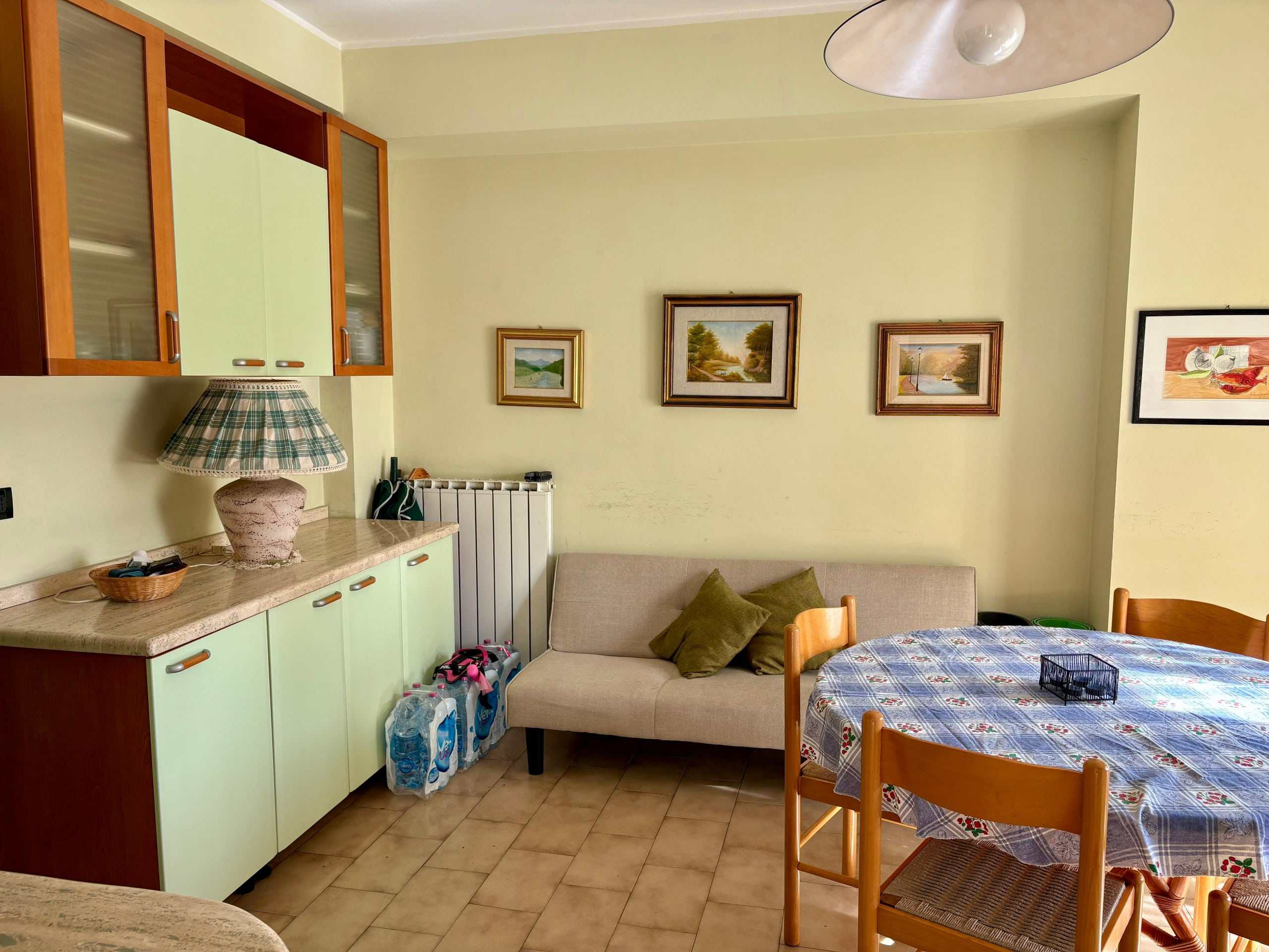 Lavanderia con mobile cucina, divano, tavolo con tovaglia e quadri alle pareti, ambiente accogliente e luminoso, arredamento pratico e funzionale, ideale per abitazioni e affitti turistici.