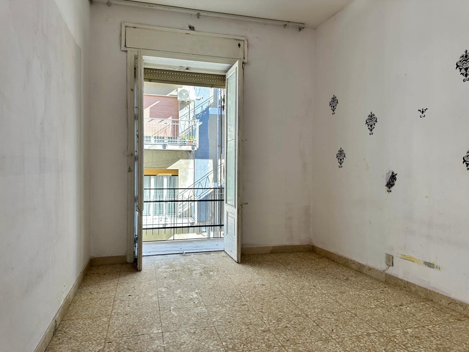 Spazio abitativo vuoto con balcone e vista su edificio vicino, pareti bianche con decorazioni nere, pavimento in piastrelle beige, ideale per ristrutturazione.