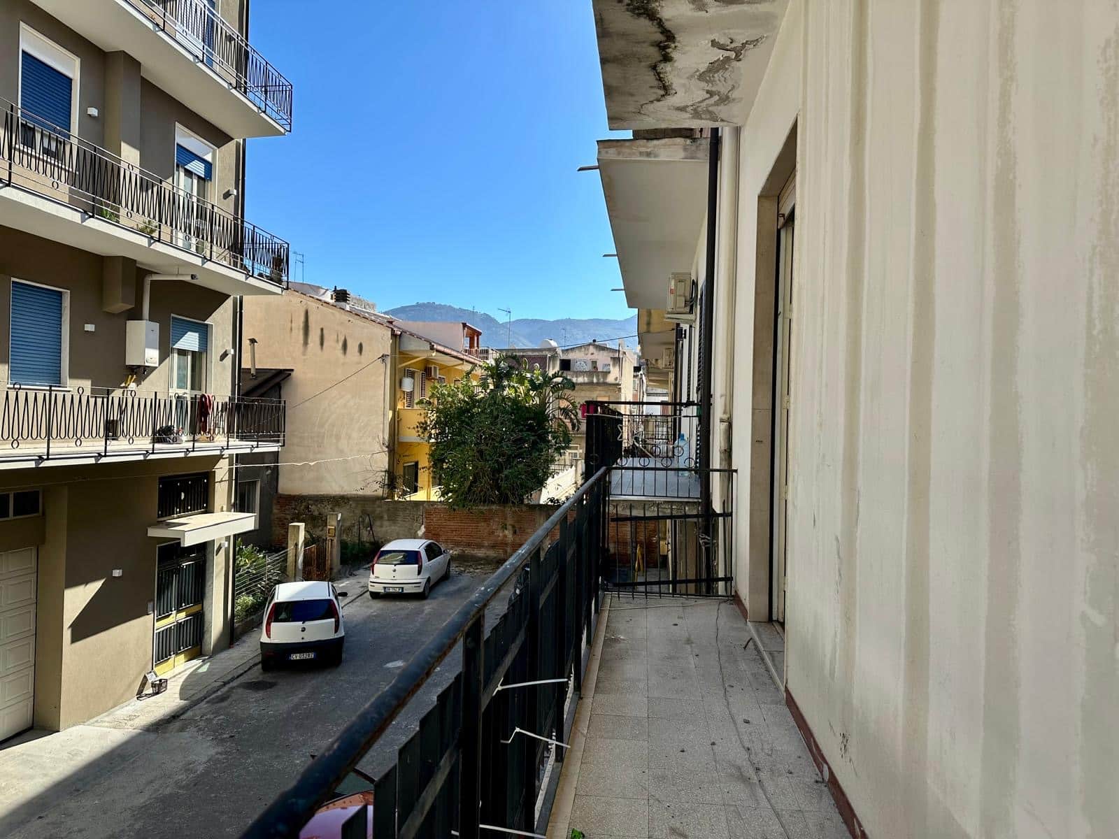 Balcone di un appartamento con vista su strada, tra edifici residenziali e montagne sullo sfondo, in un quartiere urbano di Napoli.