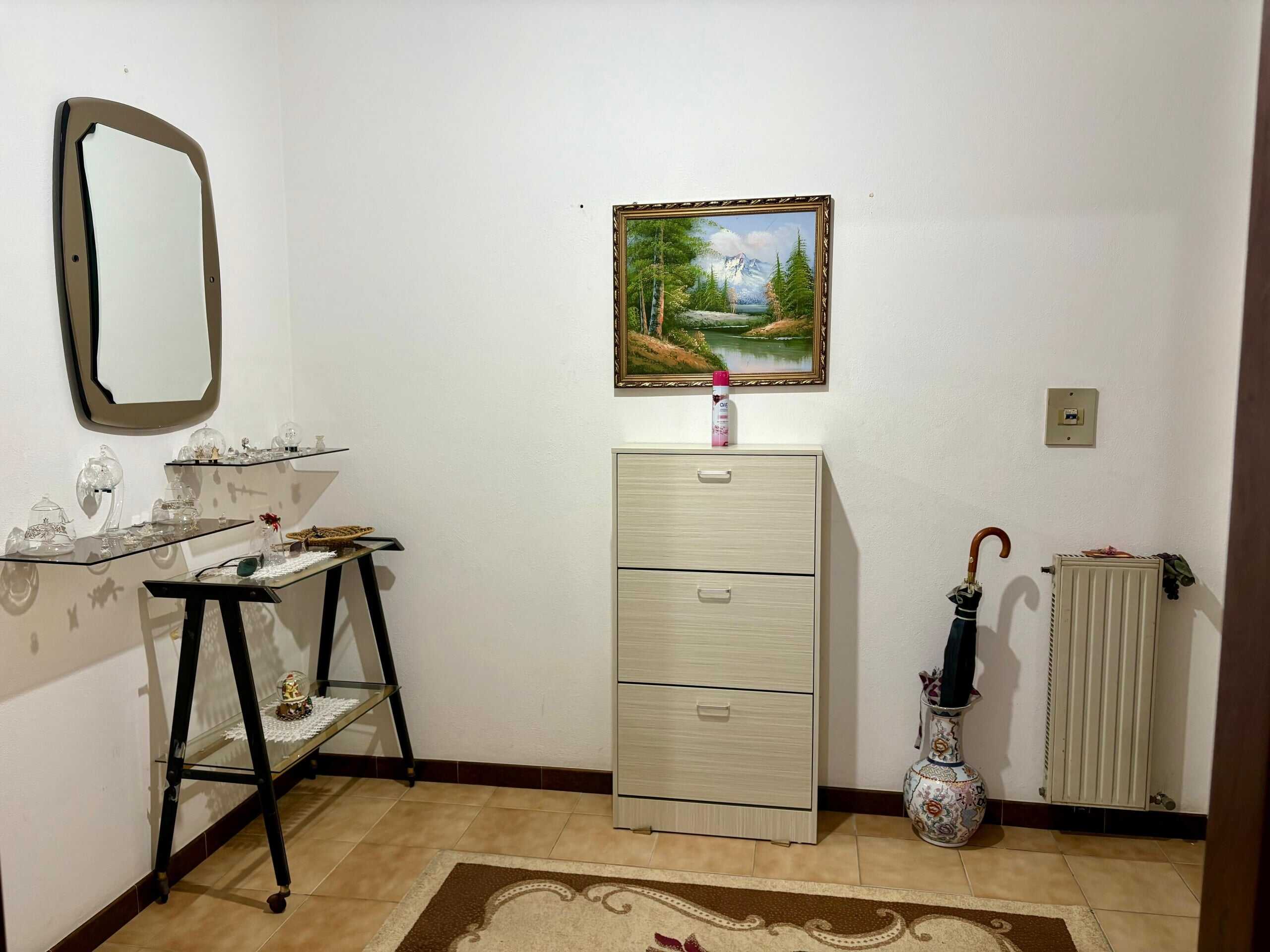 Elegante ingresso di casa con mobile a due ante, parete decorata con dipinto paesaggistico, specchio a parete e poggiapiedi con accessori decorativi, atmosfera accogliente e ben curata.