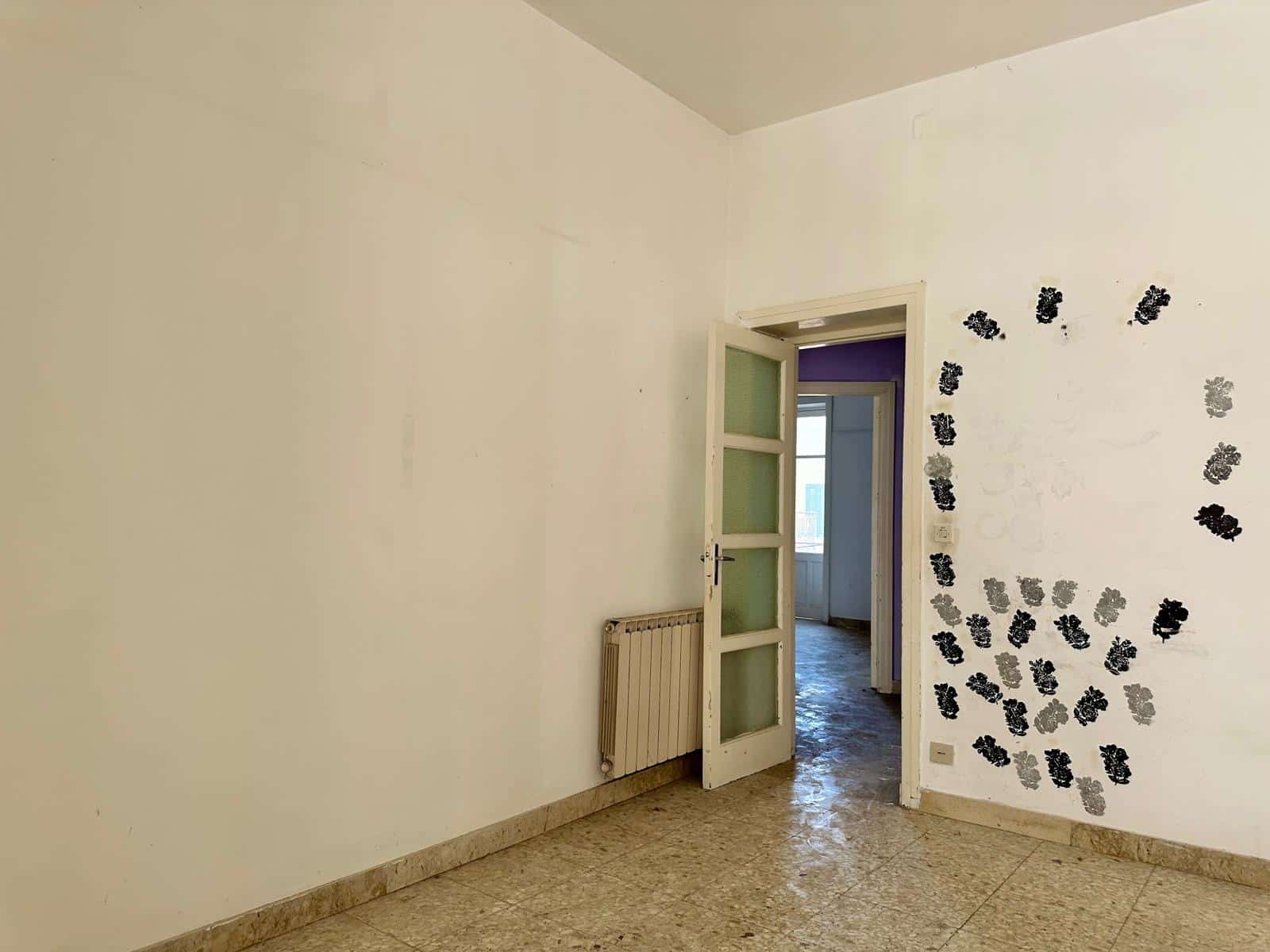Notte di una stanza vuota con pareti bianche e un pavimento in marmo, porta aperta che conduce a un'altra stanza con pareti viola e una finestra. Decorazioni con forme di foglie nere e grigie sulla parete a destra.