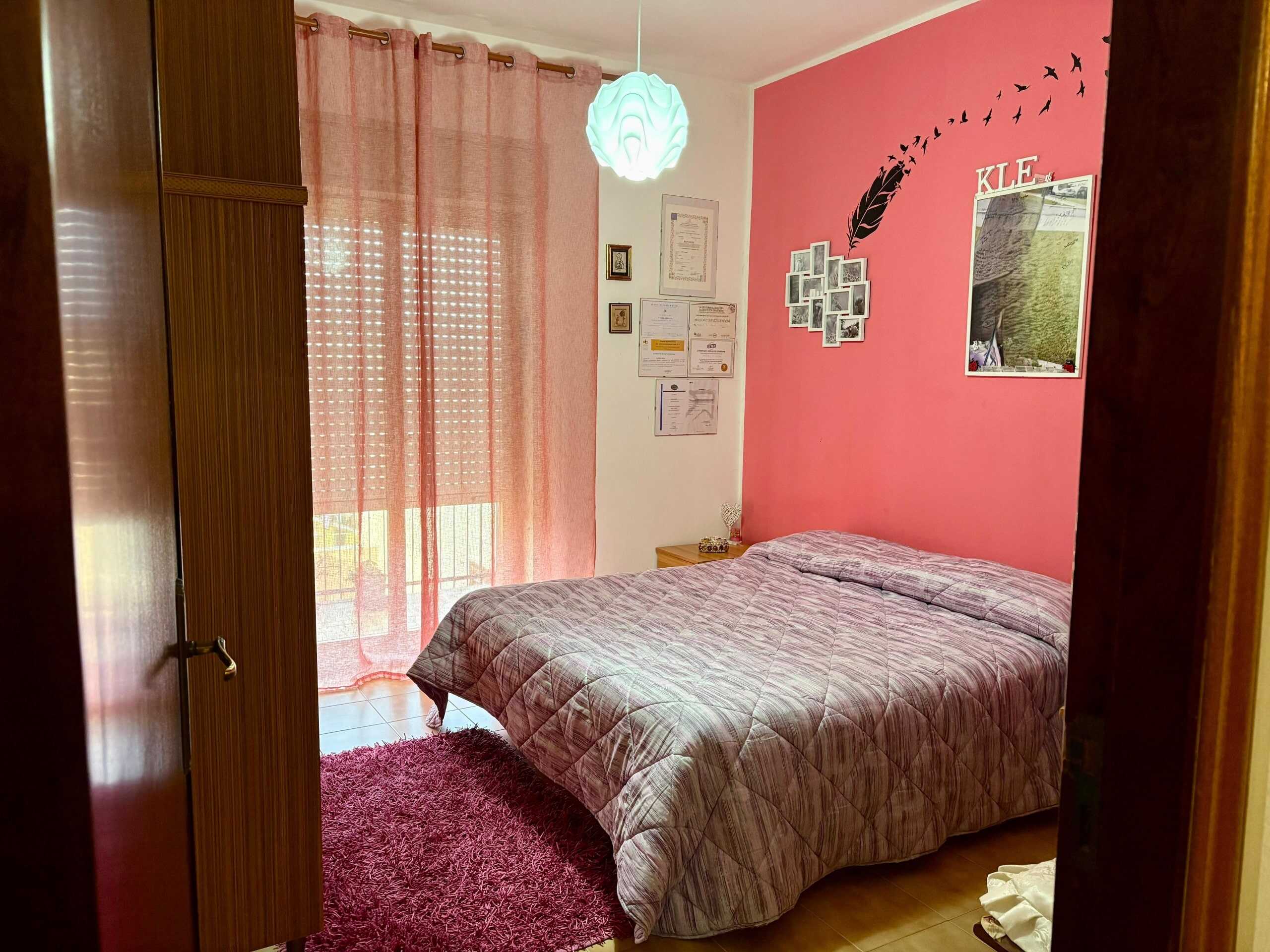 Letto matrimoniale con copertura a quadri e pareti color rosa, arredamento accogliente e pareti decorate con foto e lettere, illuminazione soffusa, ambiente ideale per camere da letto confortevoli e moderne.