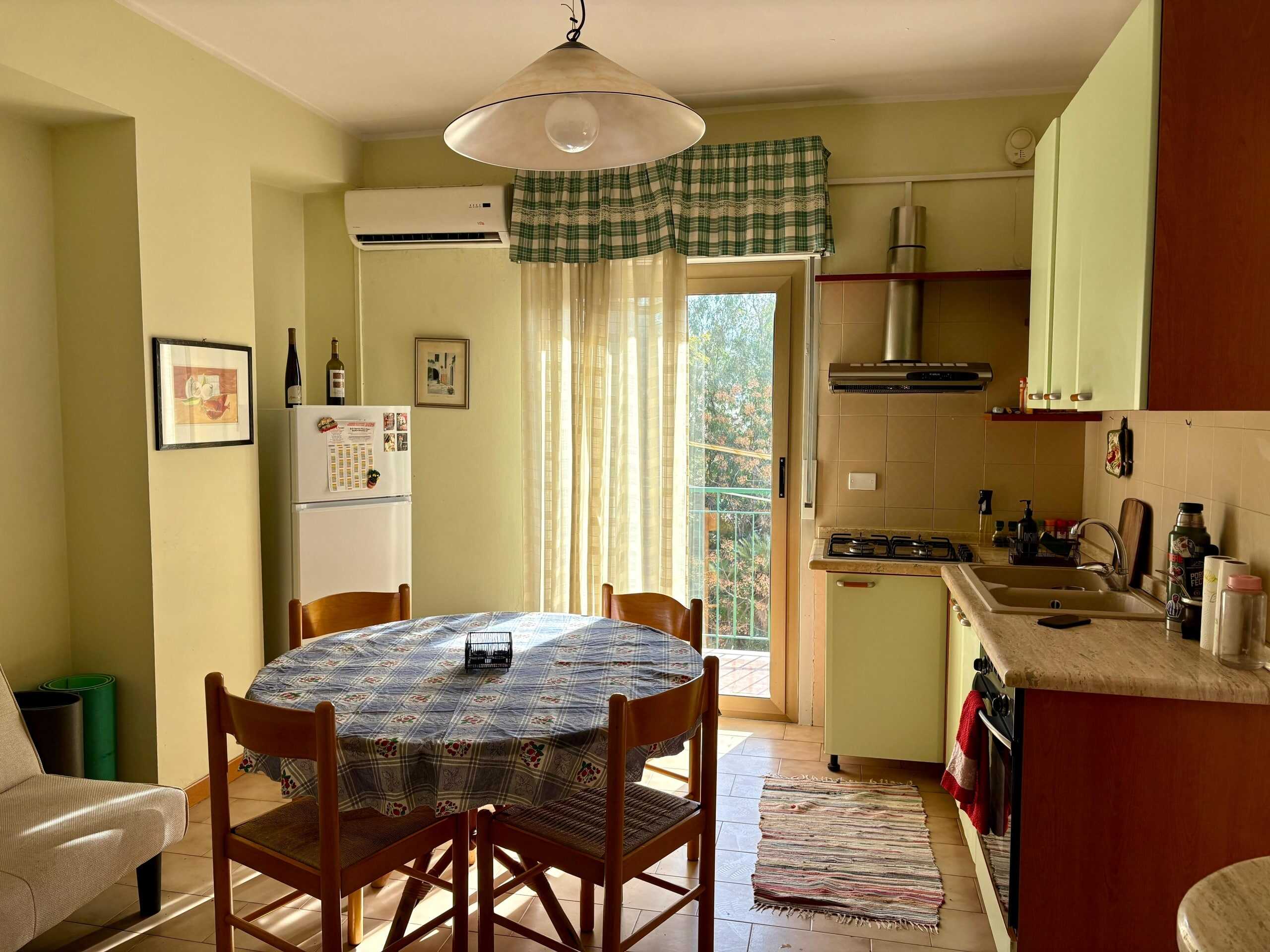 Finestra sulla balconata in cucina moderna con illuminazione naturale, arredamento accogliente con tavolo rotondo, sedie di legno, zona cottura con piano cottura e accessori, atmosfera calda e funzionale.