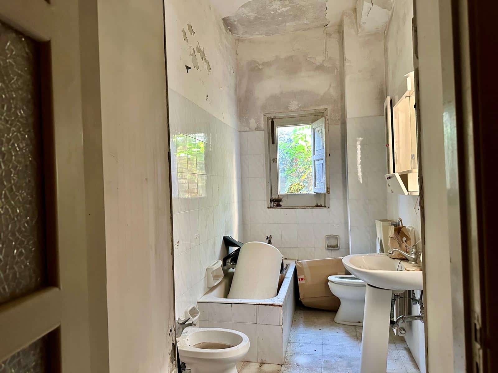 Bagno da ristrutturare con vasca, lavabo e WC, finestra che dà sulla natura, pareti e soffitti in fase di restauro, ideale per progetto di ristrutturazione o rinnovamento abitativo.