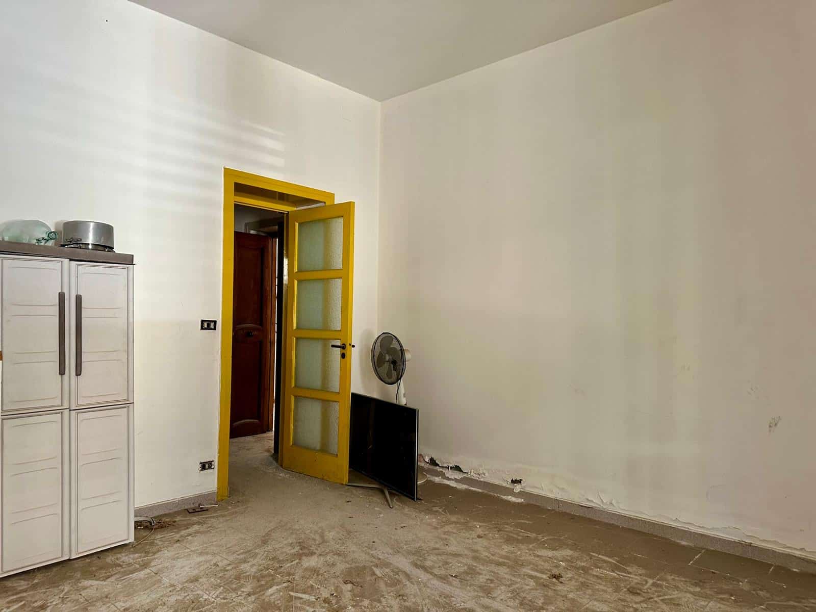 Rustico da ristrutturare con pareti bianche, pavimento in cemento grezzo, porta gialla e finestra con vetri opachi, spazio interno con mobili essenziali, ideale per investimento immobiliare.