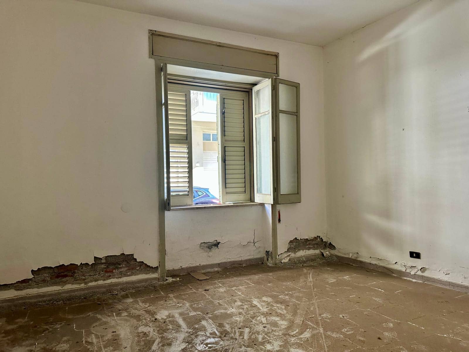 Finestra con persiane bianche in una stanza in fase di ristrutturazione, pareti con danni e pavimento in cemento. Ideale per interventi di ristrutturazione immobiliare e ristrutturazioni edilizie.