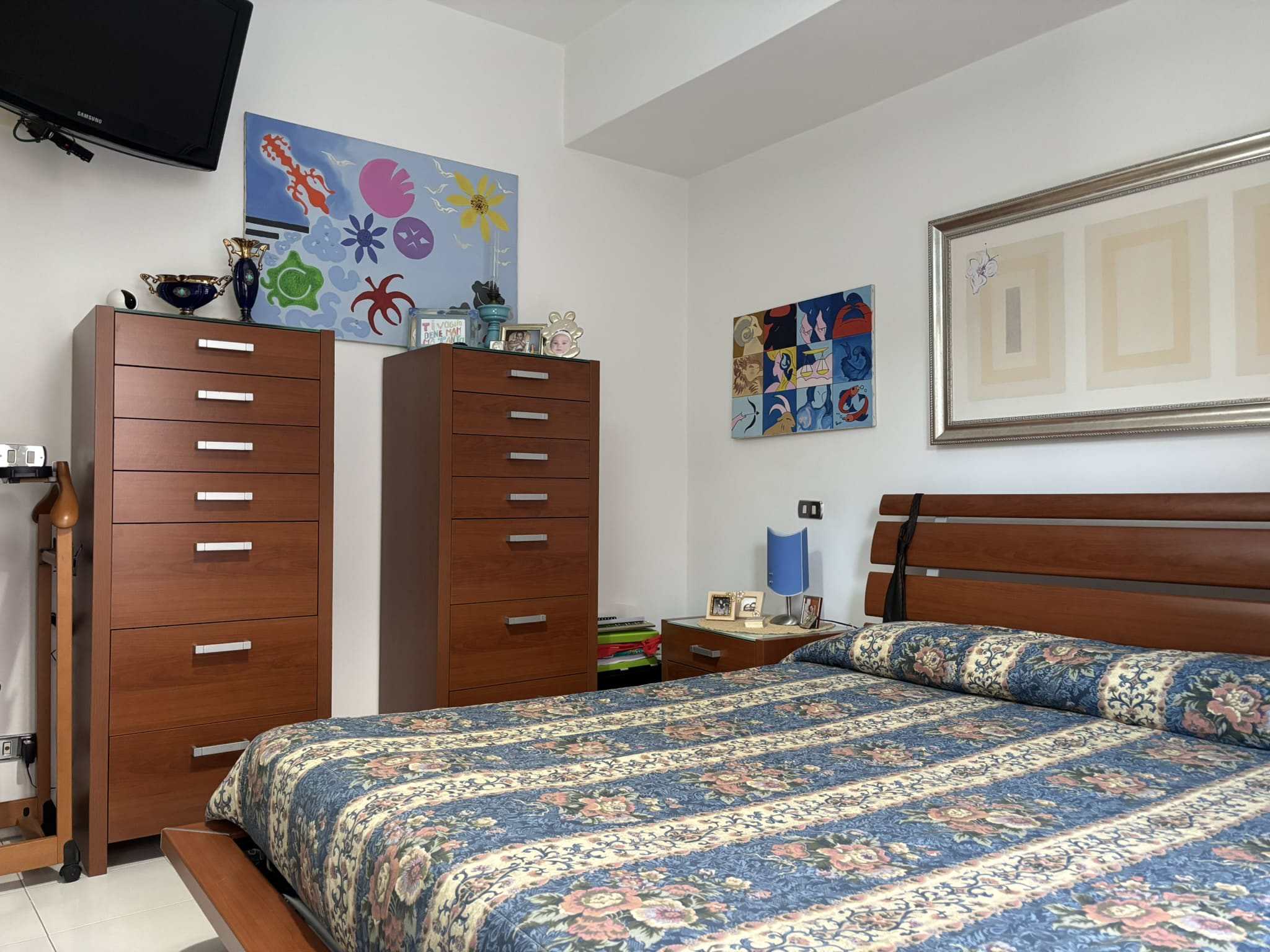 Armadio in legno con comò in camera da letto, decorata con opere d'arte colorate e oggetti personali, ambientazione di stile moderno e accogliente per l'arredamento di case e appartamenti.