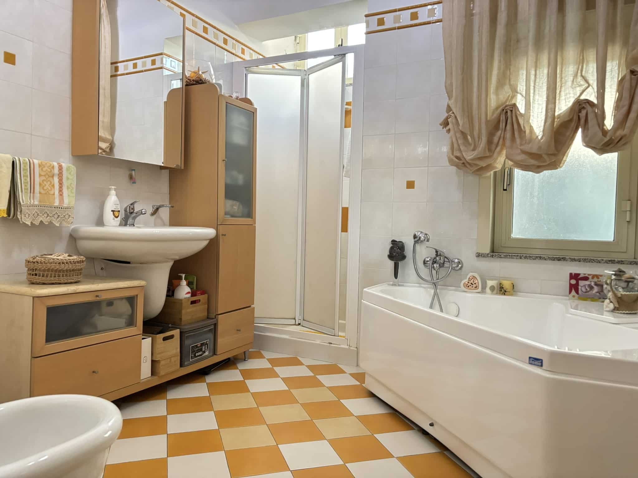 Bagno spazioso con vasca, doccia e arredi in legno chiaro, finestra con tende beige, piastrelle colorate in stile vintage, ideale per casa o appartamento.