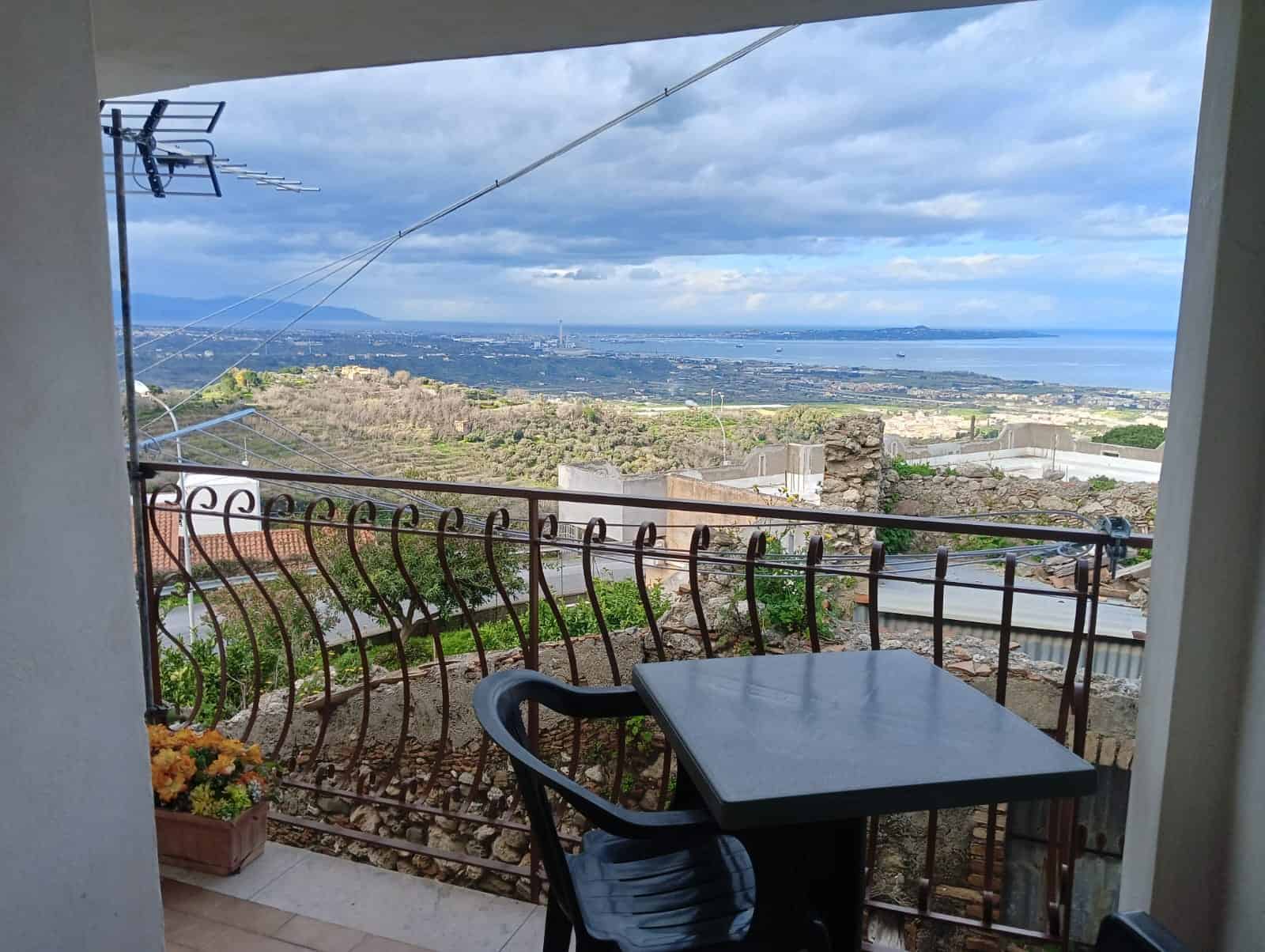 123c5bf6-374d-4355-b1eb-43aaf67cc27b Vista panoramica mare e colline da terrazza con balcone in stile rustico, arredato con tavolo e sedie, su sfondo di cielo nuvoloso, suggestivo scenario di appartamento NextCasa Italia.