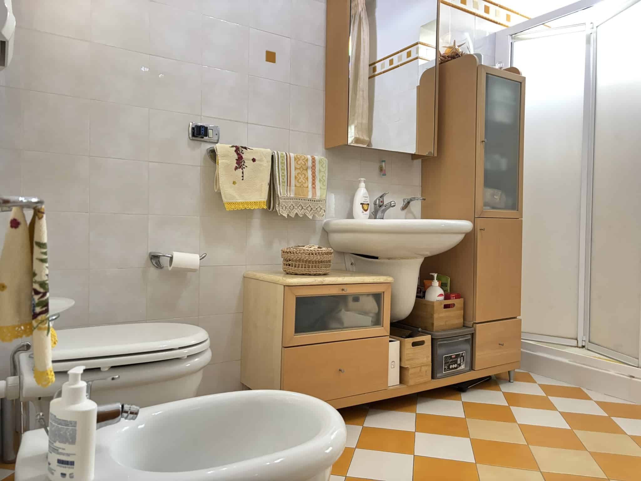 Lavabo e mobili bagno in legno chiaro con specchi e accessori, pavimento a losanghe in ceramica beige e arancione, box doccia e strumenti di uso quotidiano, in un bagno accogliente e funzionale.