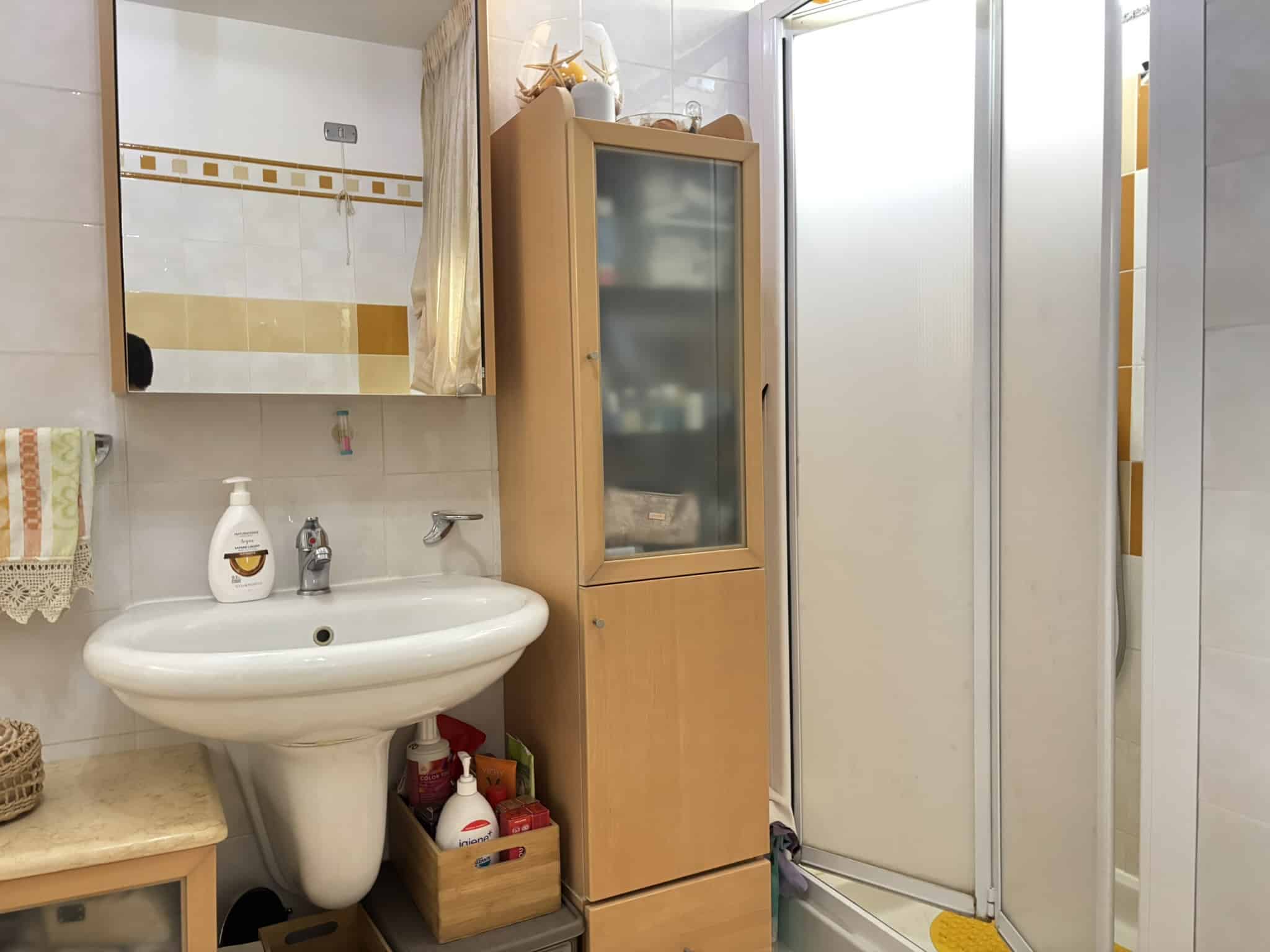 Locale bagno con lavabo, specchio, mobiletto in legno, cabina doccia e vari detergenti, arredamento funzionale, spazio ben organizzato, stile pratico e accogliente.