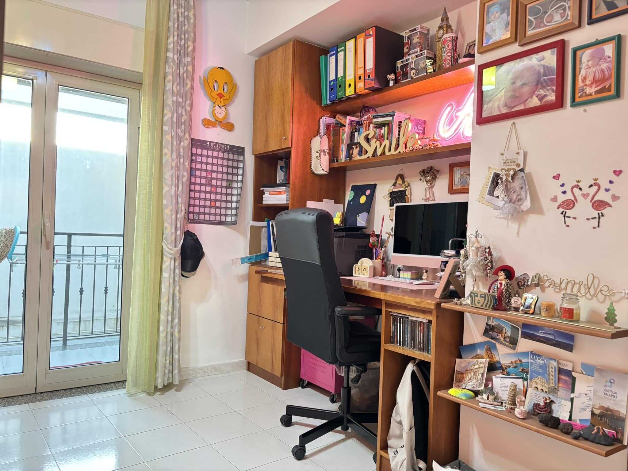 Scrivania da ufficio in casa con album, libri e decorazioni, vicino a balcone con tende leggere, ambiente luminoso e accogliente, ideale per smart working e studio creativo.