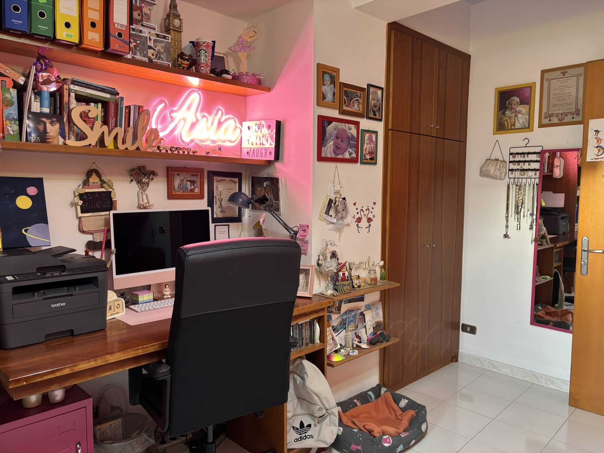 Scrivania con computer e stampante, pareti decorate con foto, quadri e luci neon rosa, armadi in legno, specchio a parete, oggetti decorativi e ripostiglio, ideale per ufficio o zona studio.