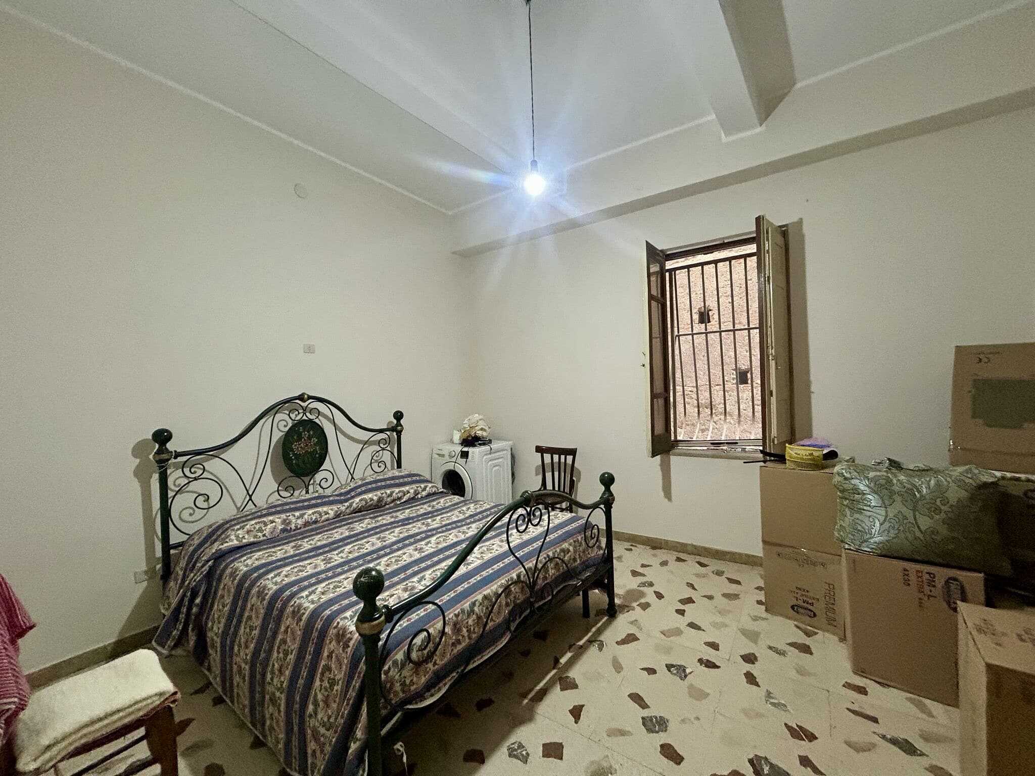 Foto di una camera da letto semplice e funzionale, con letto in ferro battuto, finestra con grate e arredamento minimale. La stanza si presenta luminosa, con pareti chiare e pavimento in pietra decorata. Perfetta per chi cerca soluzioni abitative pratiche e di valore.