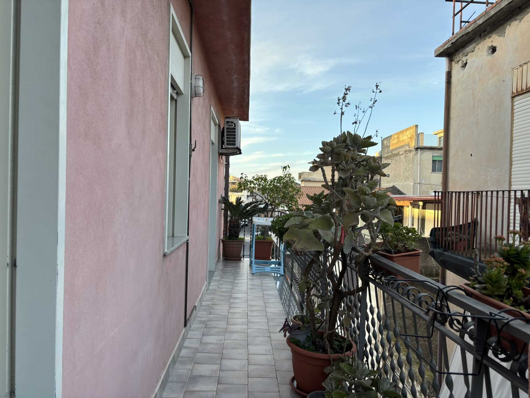 Balcone di un appartamento con piante in vasi, vista sulla città, con pareti esterne color rosa e pavimentazione in gres porcellanato, ambiente accogliente e luminoso.