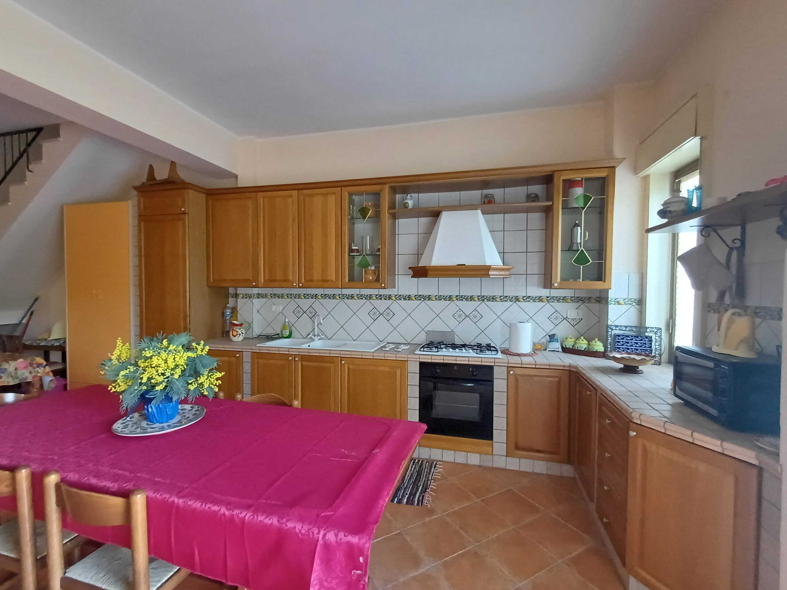Voliera cucina in stile rustico con mobili in legno, tavolo con copertura rosa e decorazioni floreali, forno a gas e accessori da cucina, grande finestra per illuminazione naturale e pareti con piastrelle decorate, ambiente accogliente e funzionale.
