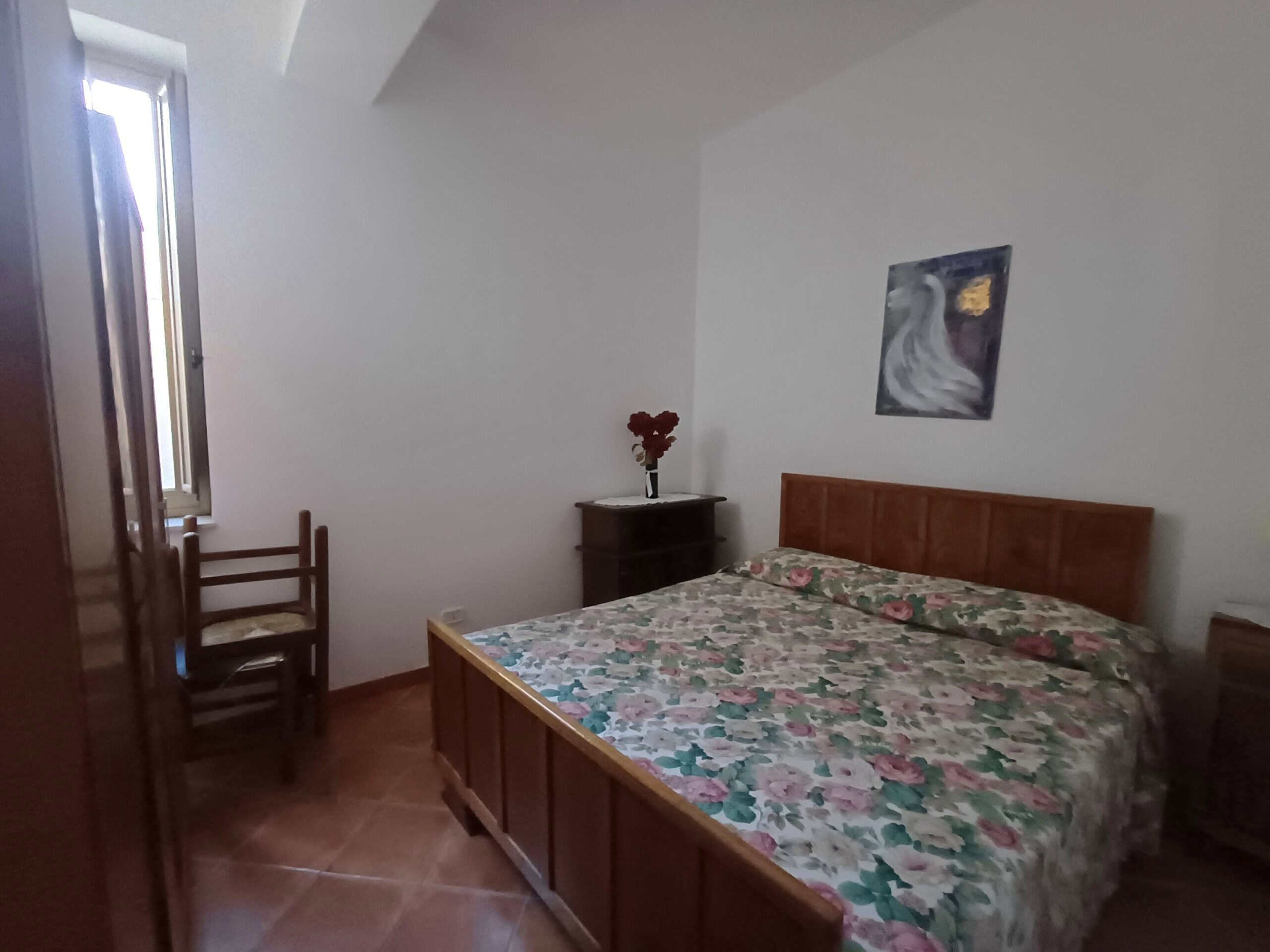 Camere da letto con arredi in legno, letto matrimoniale con copriletto floreale, finestra con veneziana, decorazioni artistiche sulla parete, ambiente accogliente e luminoso.