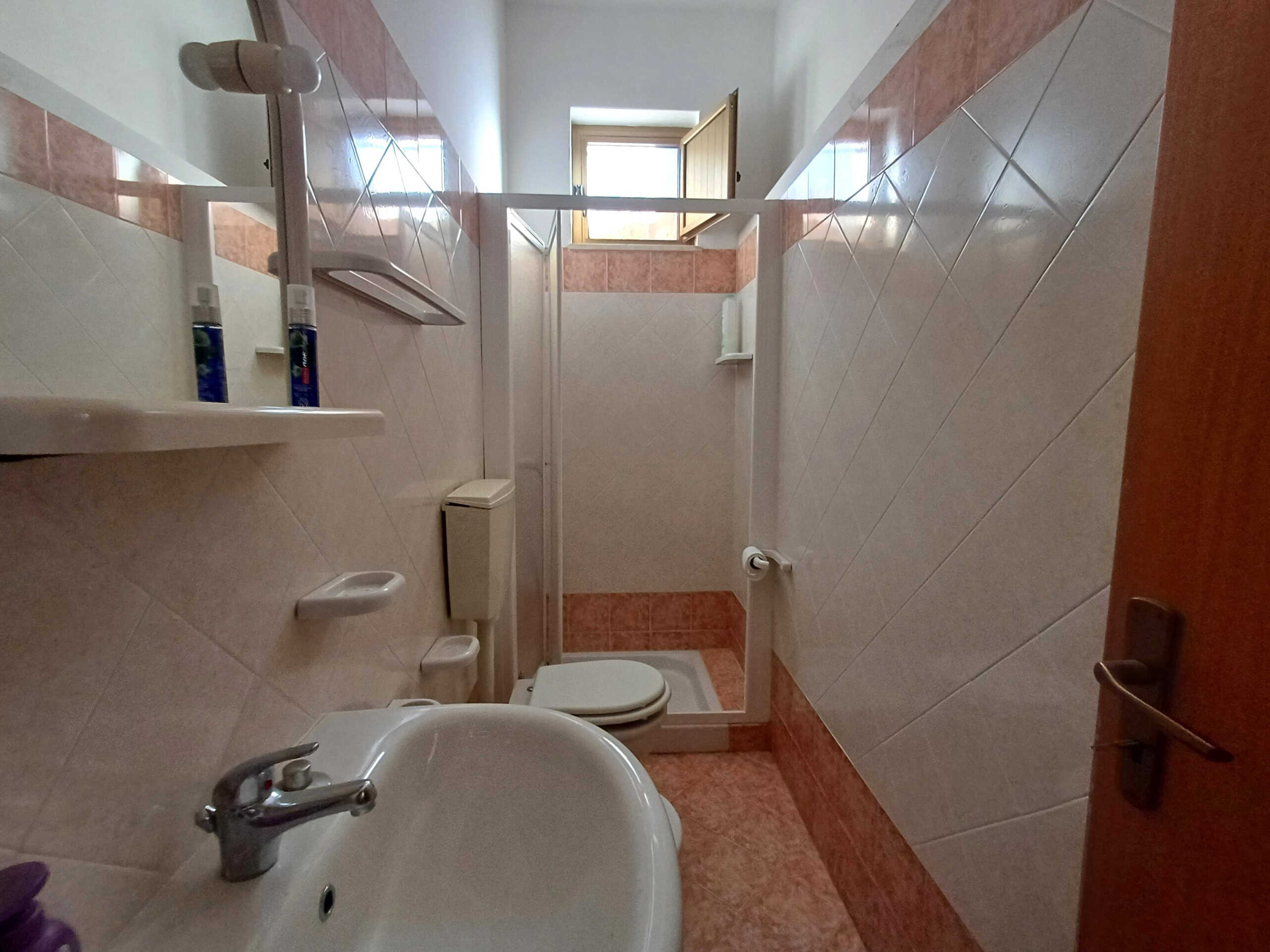 Lavabo e servizi igienici in un bagno di piccole dimensioni, con piastrelle rosa e bianche, finestra che permette l'ingresso di luce naturale, ideale per appartamenti vicini ai servizi di prima necessità.