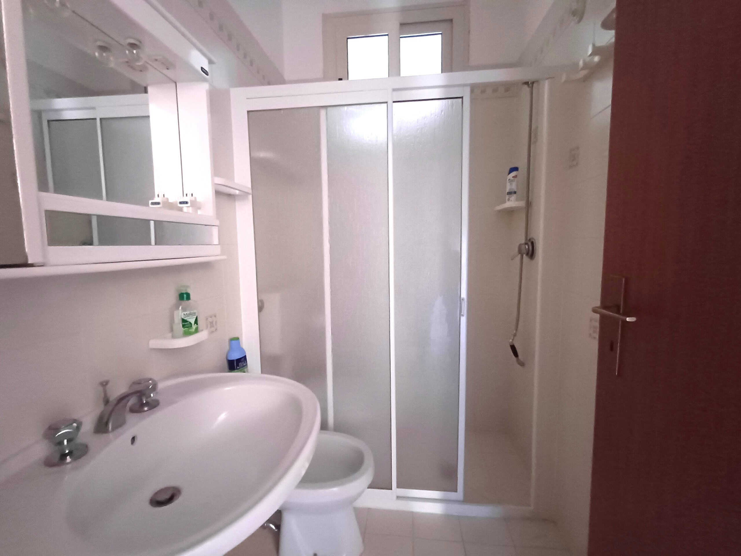 Doccia in bagno con pannelli in vetro, lavabo bianco con accessori, tendo doccia, e scaffale portaoggetti in vetro - Ideale per soluzioni di ristrutturazione bagno moderne e funzionali.