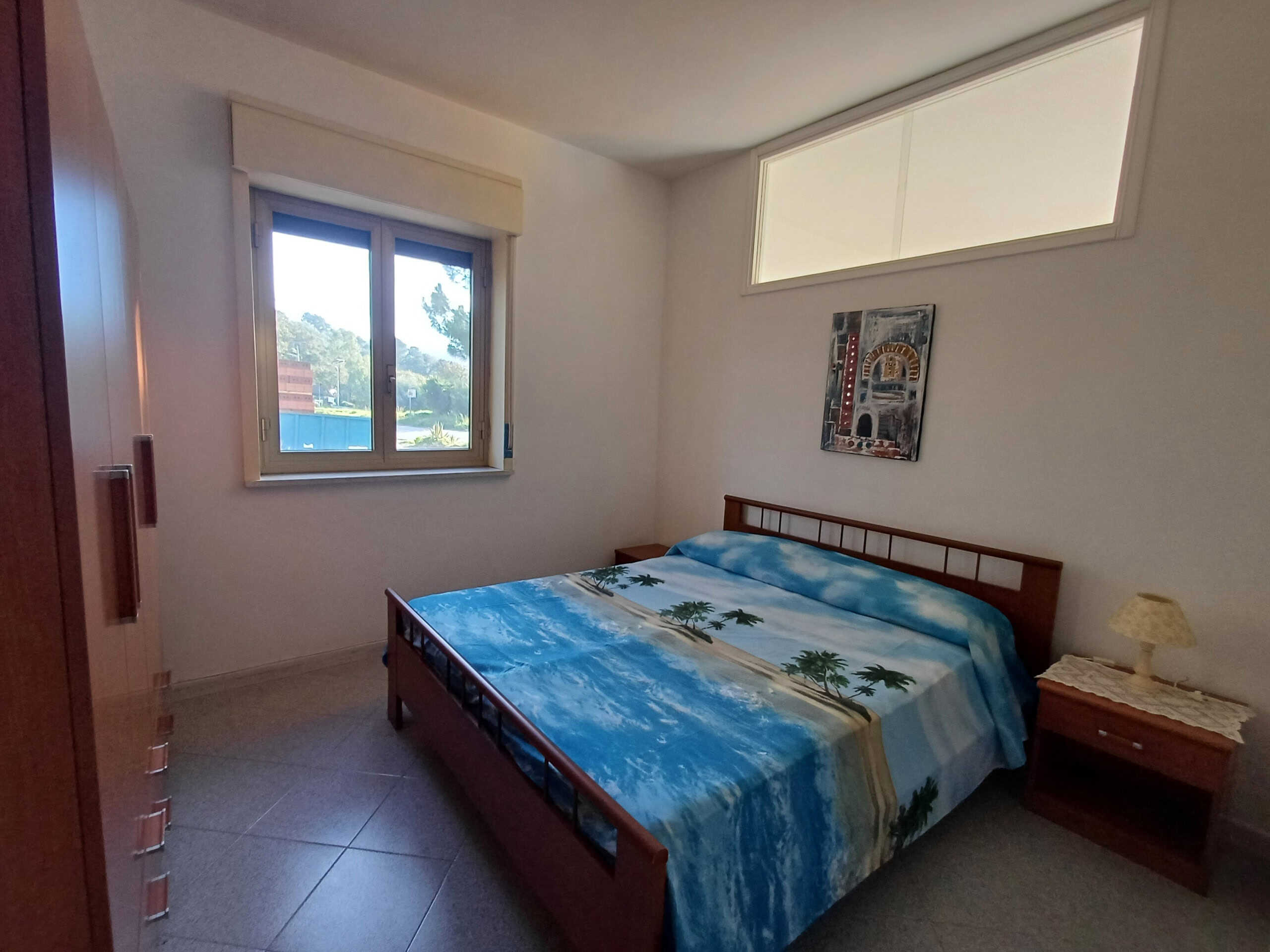 Fotografia di una camera da letto luminosa con letto matrimoniale, decorata con biancheria a tema spiaggia, finestre con vista sul verde e mobili in legno, ambiente confortevole e accogliente, ideale per case vacanze o residenze al mare.