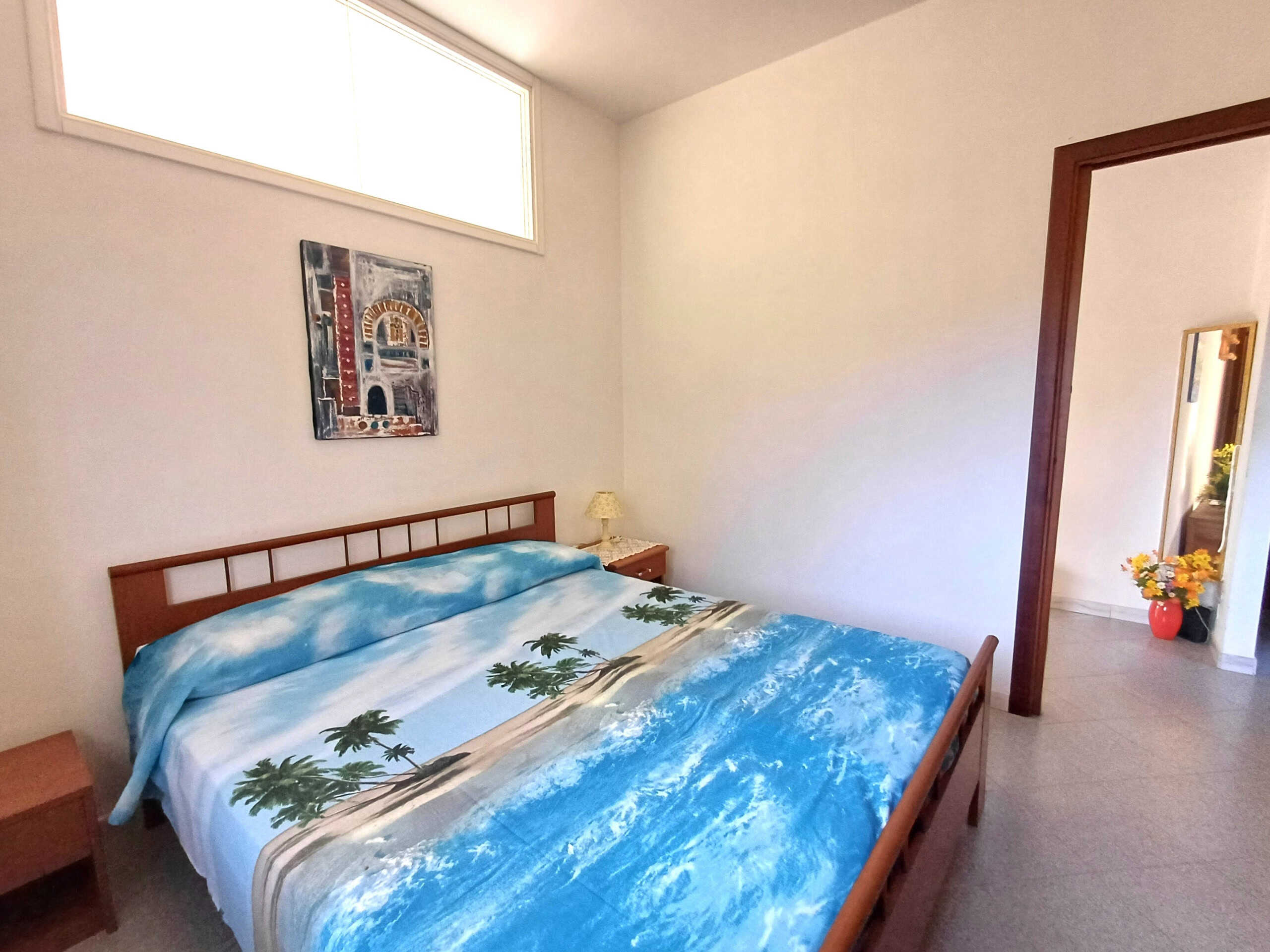 Letto matrimoniale con coperta a tema mare e palme, stanza luminosa con pareti bianche e finestra alta, arredi in legno, decorazioni artistiche e ambiente accogliente.