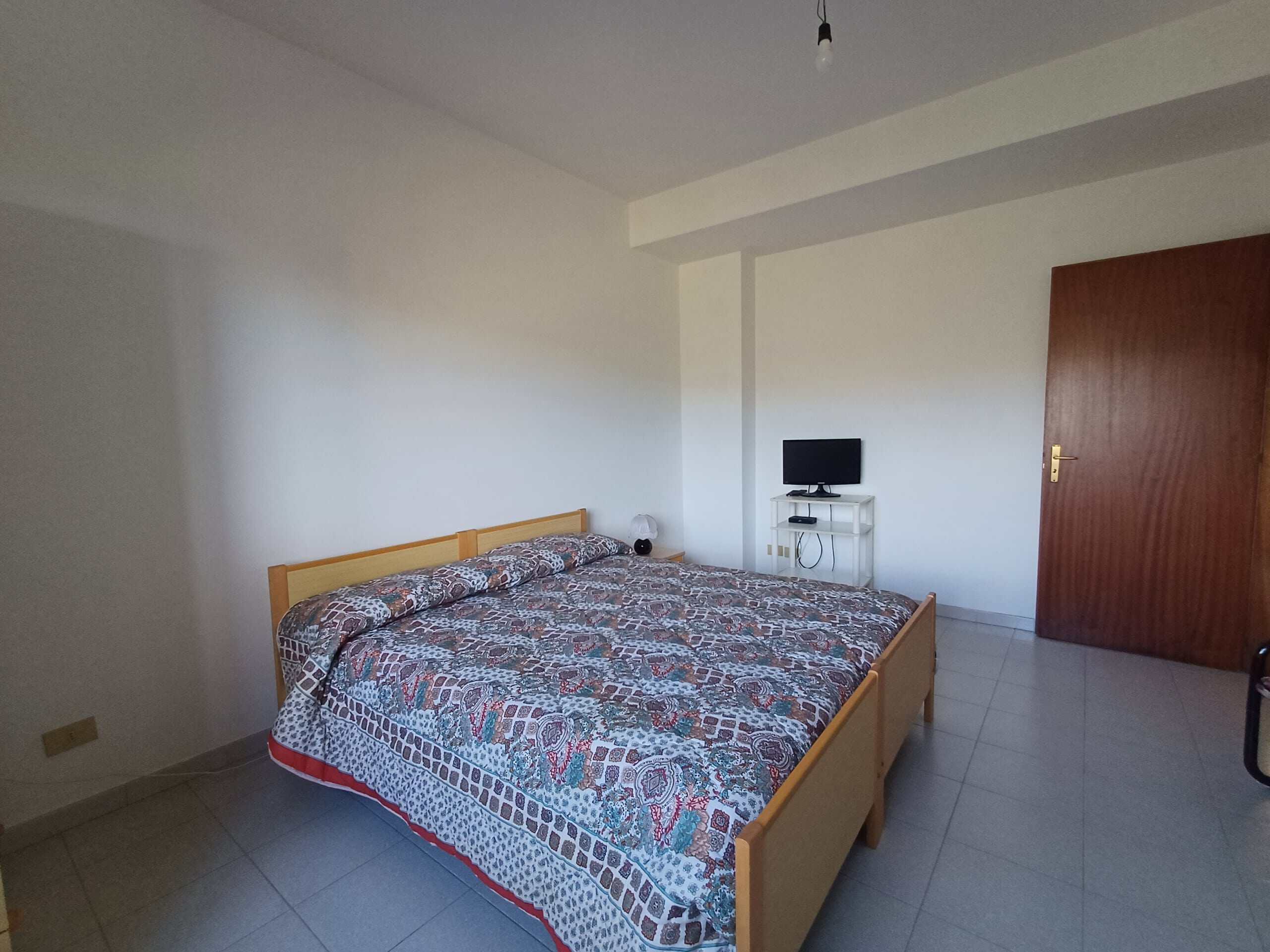 Letto matrimoniale con biancheria colorata in camera da letto spaziosa e luminosa con TV moderna e armadio in legno chiaro.