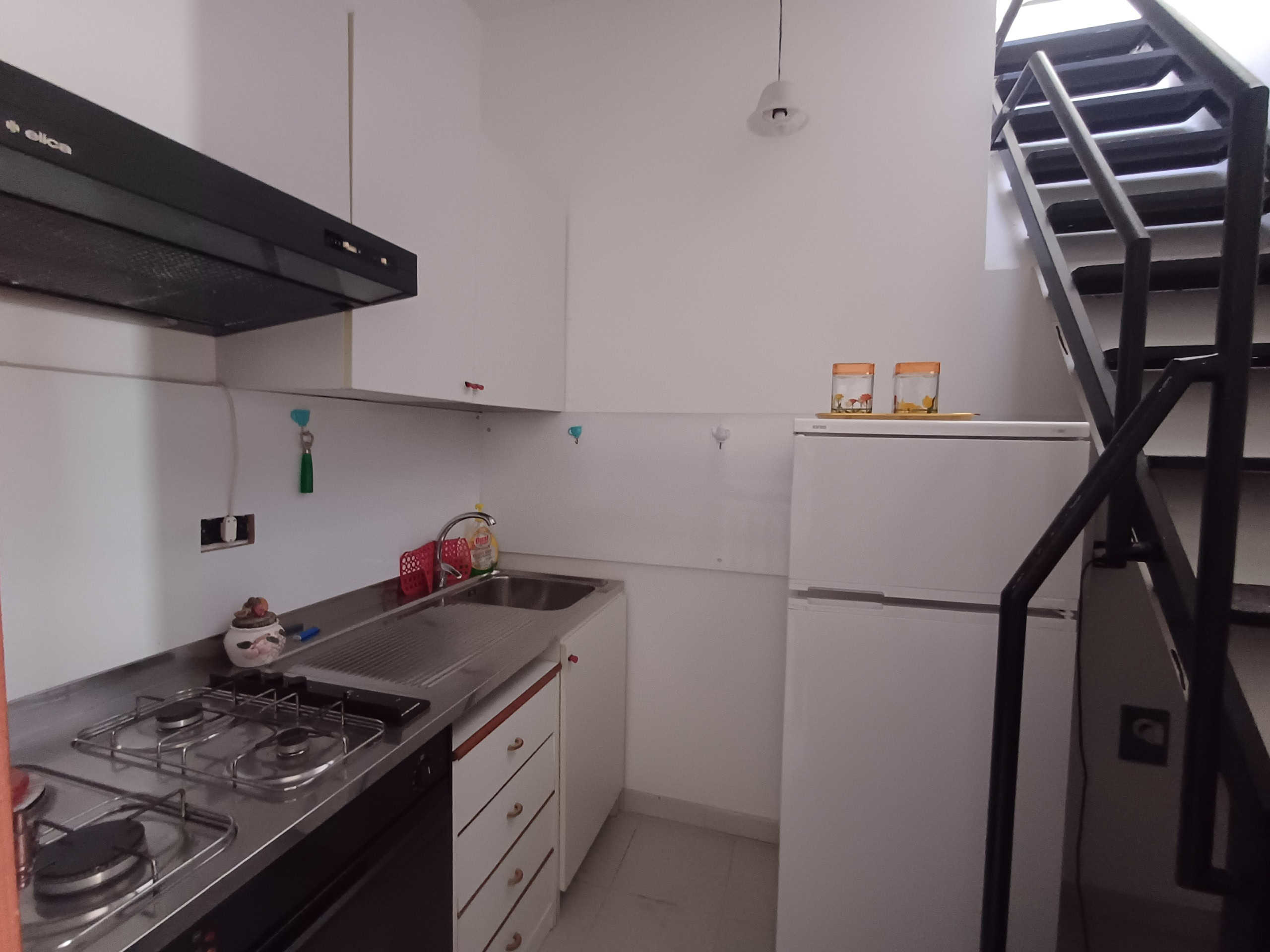 Lavanderia compatta con piano cottura, frigorifero e scale metalliche in un appartamento moderno, ideale per soluzioni abitative funzionali e ottimizzate.
