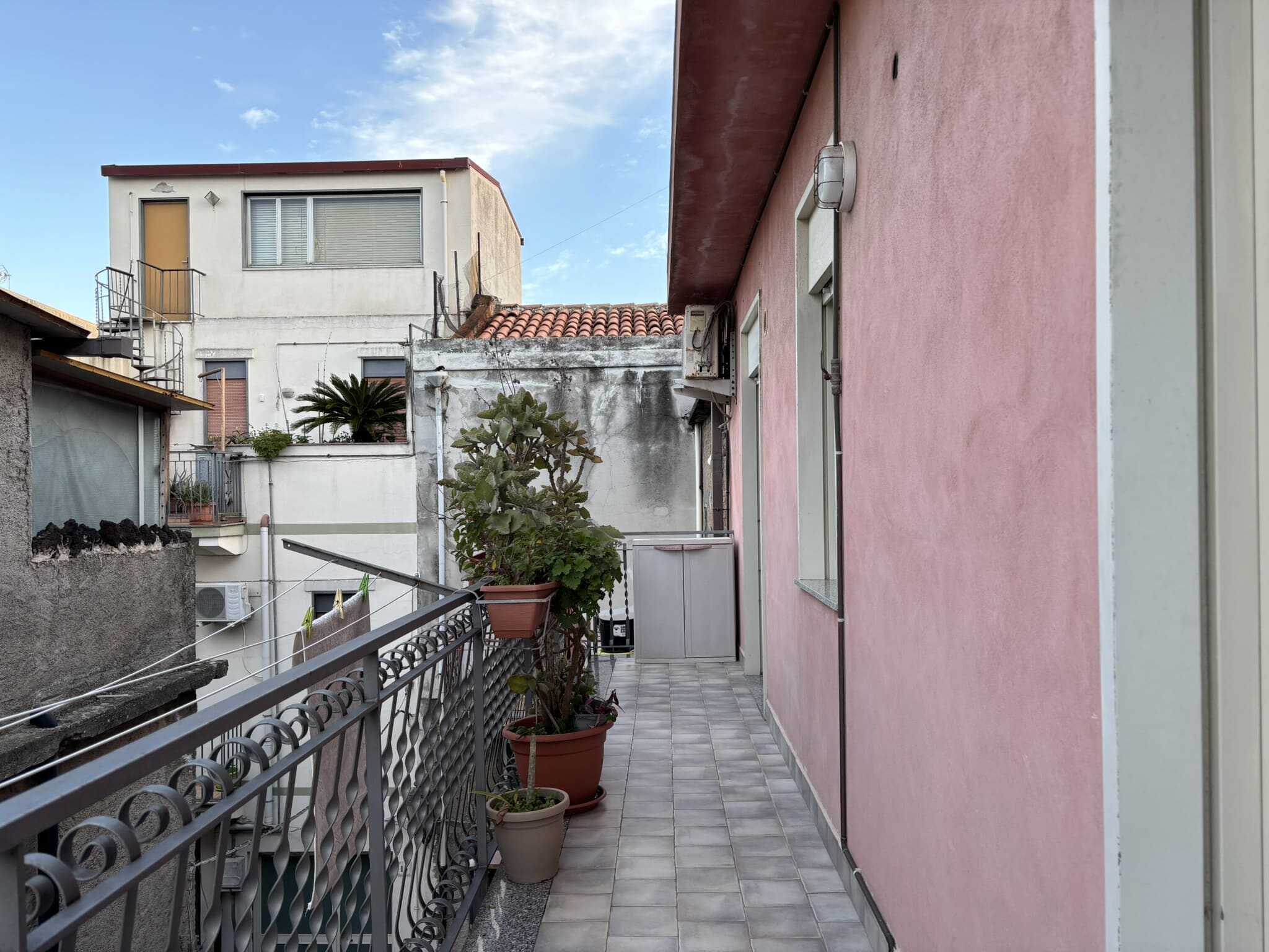 Balcone di appartamento con piante in vaso, fino a un'unità abitativa a livello strada, vista su edifici residenziali in un quartiere urbano, con cielo blu e nuvole leggere.