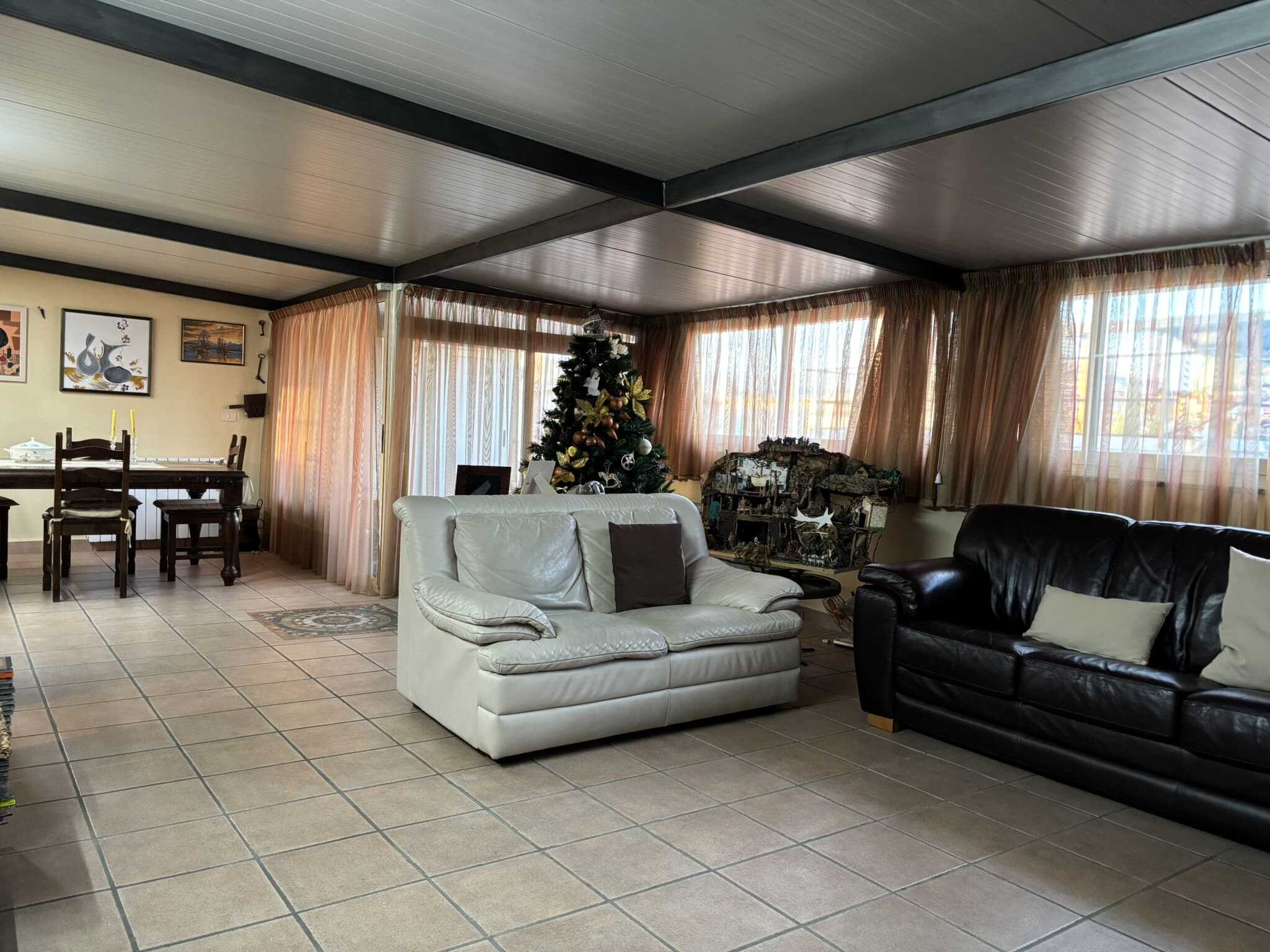 Accogliente soggiorno con divani in pelle bianca e nera, decorato per il Natale con albero decorato, ampie finestre con tende di tessuto leggero, pareti decorate con quadri e figura di presepe in miniatura.