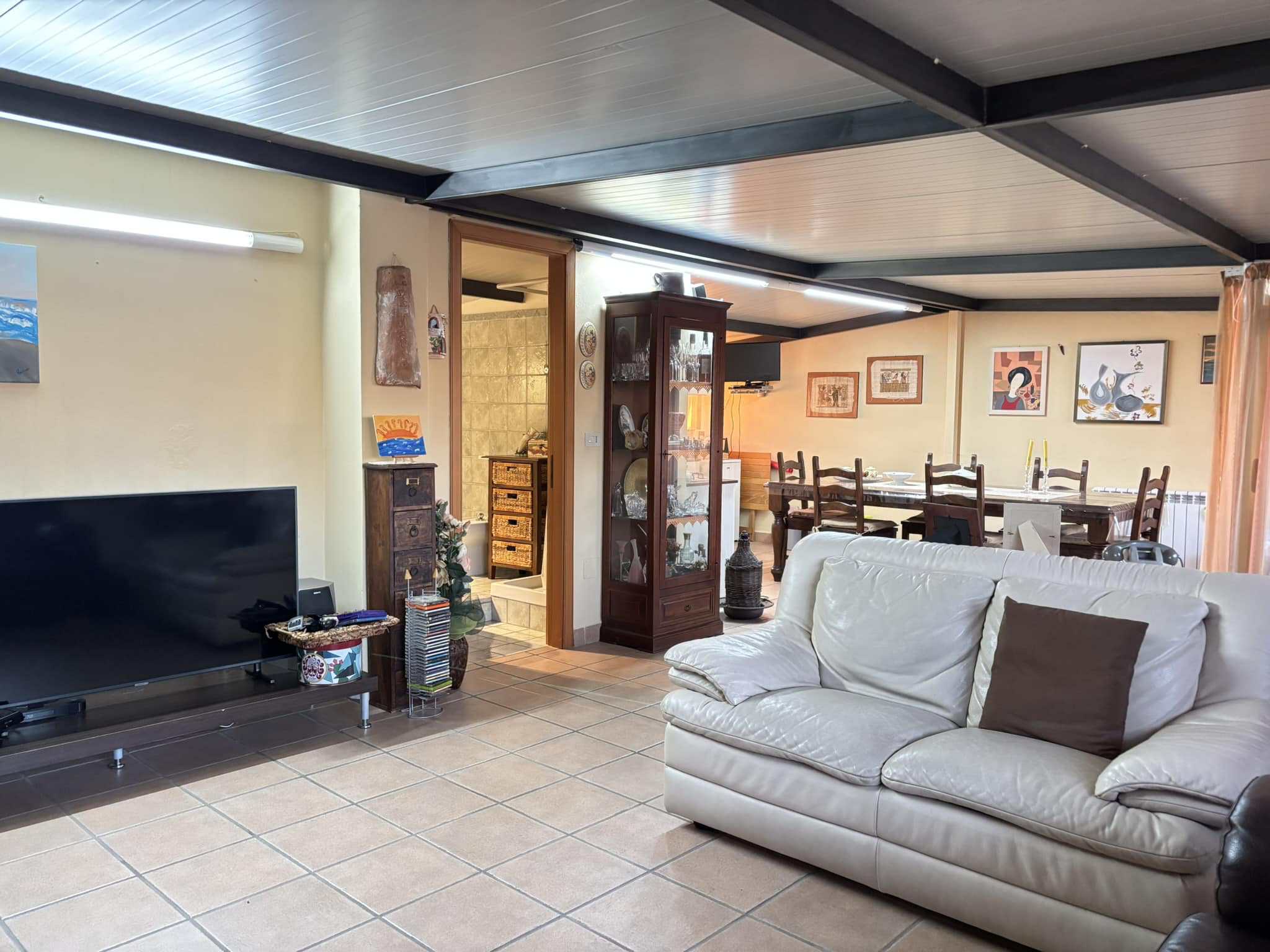 Relaxing living room with a white leather sofa, flat-screen TV, and decorative artwork, leading to a spacious dining area with wooden chairs and a table, ideal for family gatherings.