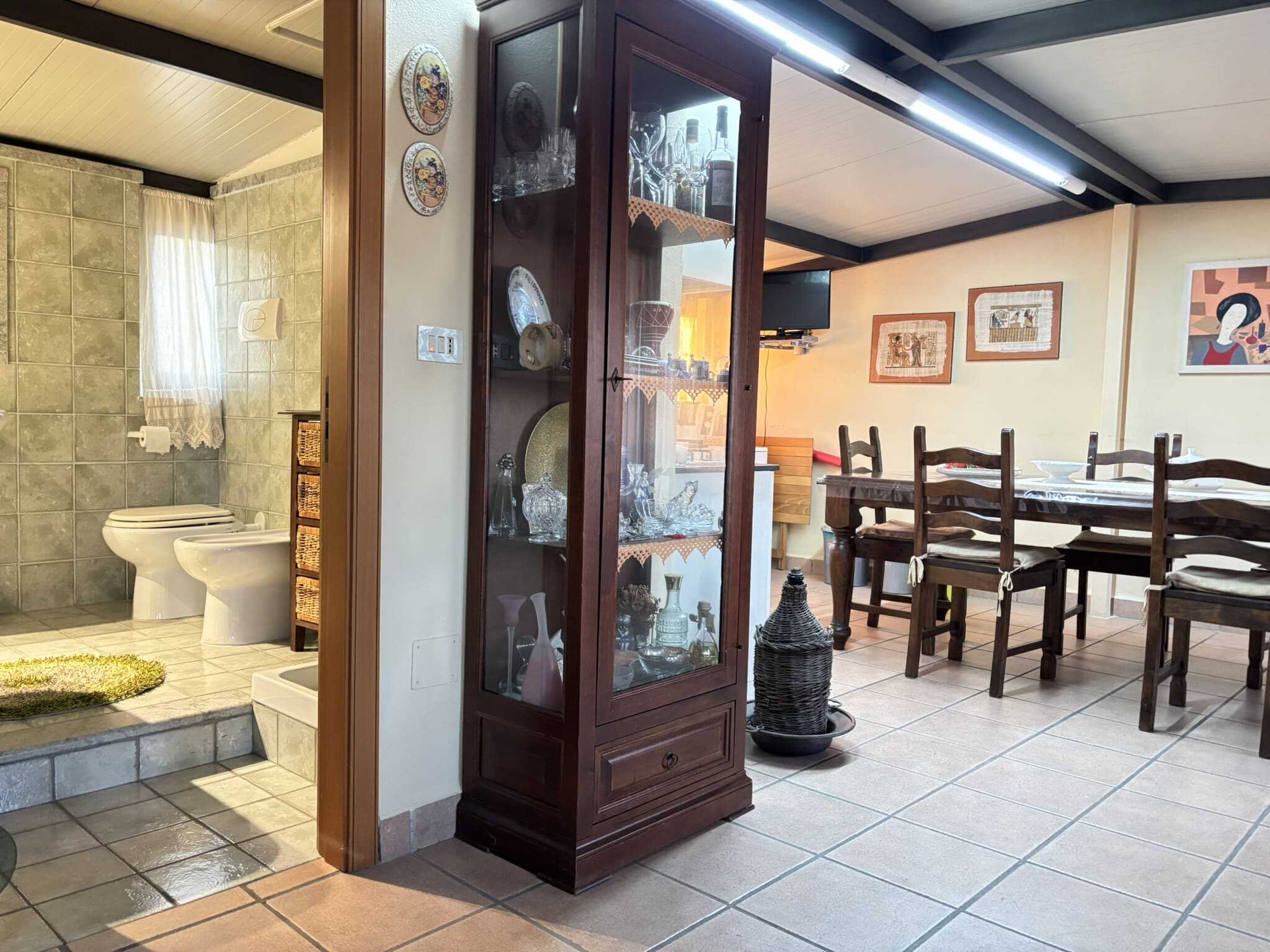 Armadio in legno con vetrina decorativa, visibile arredamento d'interni di casa con zona pranzo e bagno in background, stile rustico ed elegante.
