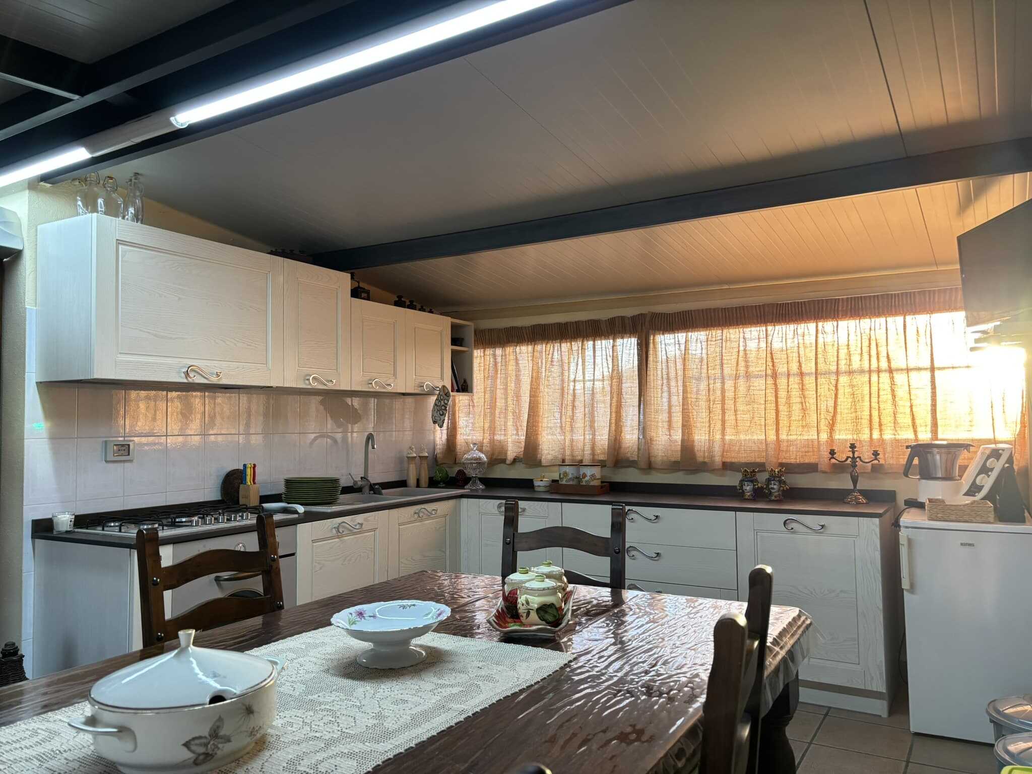 Ampia cucina luminosa con ante in legno chiaro, spaziosa area pranzo con tavolo di legno e sedie, grandi finestra con tende in tessuto che lascia passare la luce calda del tramonto, elettrodomestici moderni.