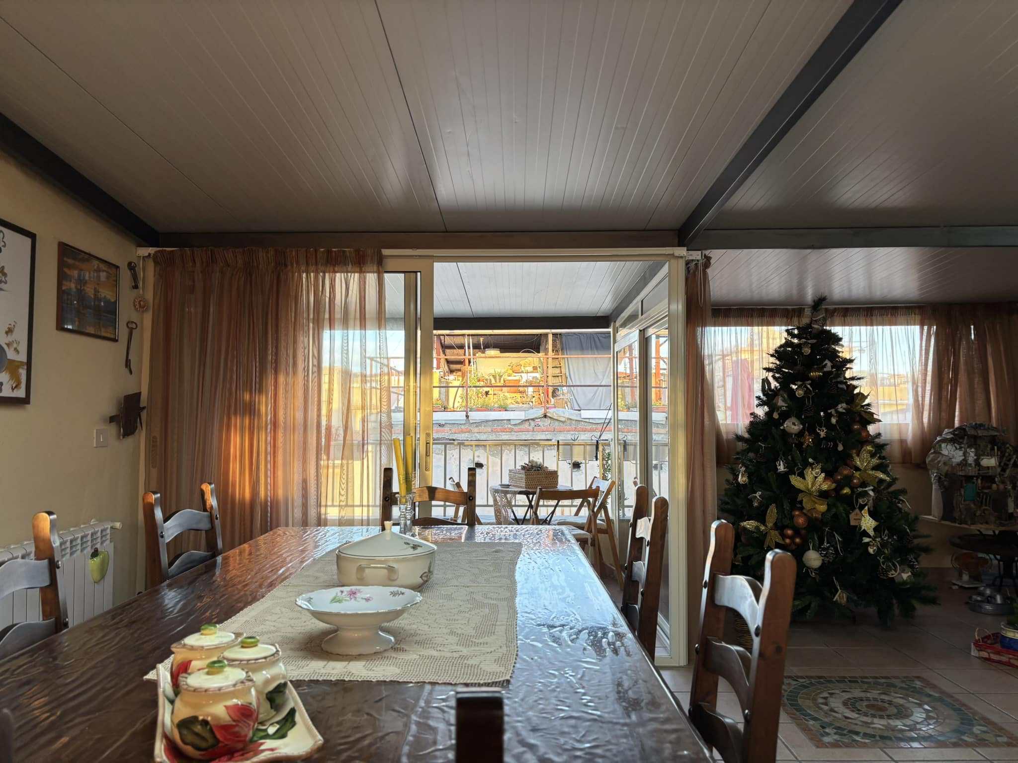 Panorama di soggiorno con tavolo da pranzo, decorazioni natalizie, albero di Natale decorato, balcone con vista luminosa, tende leggere e atmosfera accogliente per le festività.