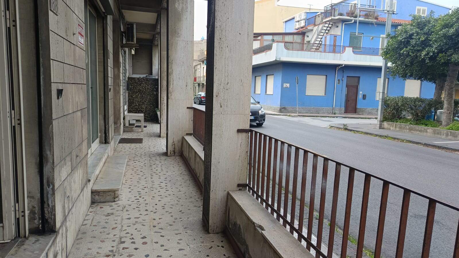 Balcone con ringhiera in metallo e vista sulla strada, edificio in stile classico, pavimentazione con piastrelle mosaico, zona residenziale urbana, case colorate e alberi, atmosfera tranquilla e silenziosa.