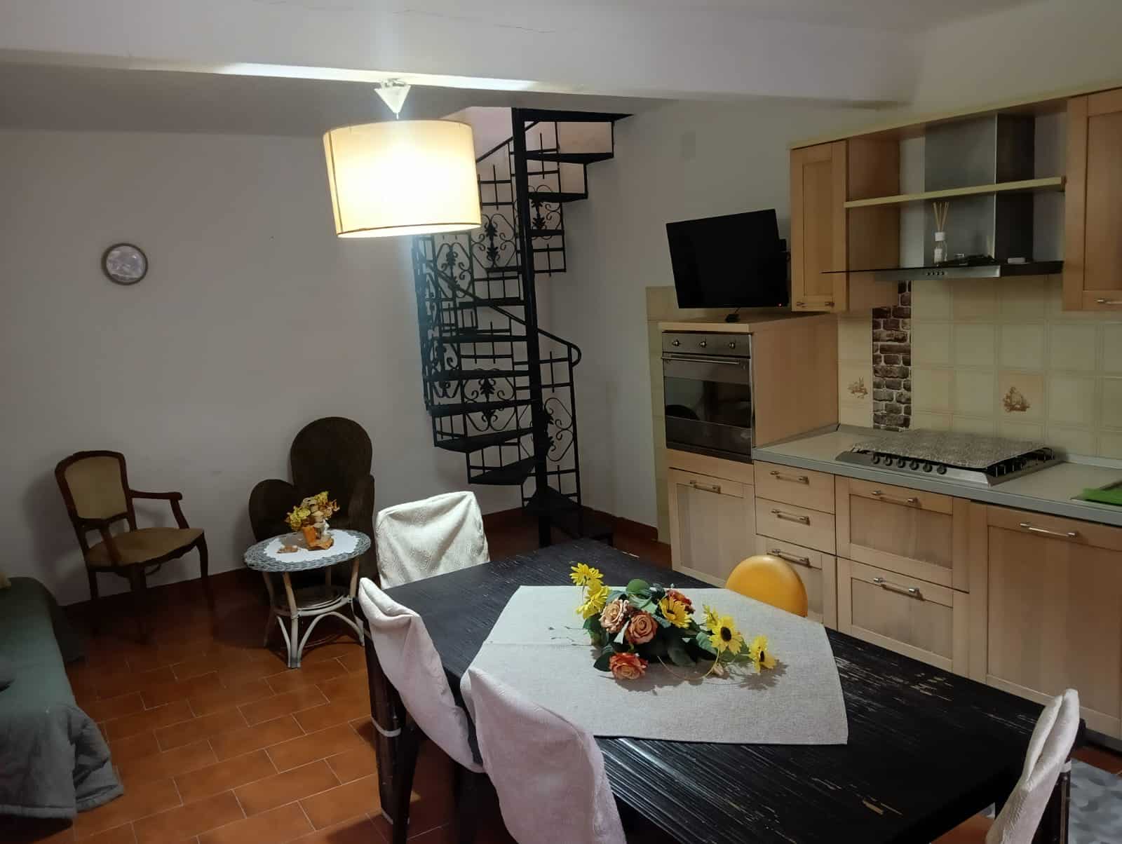 Luminoso soggiorno con cucina, tavolo da pranzo con centroflauna di fiori, sedie imbottite, scale a chiocciola in ferro battuto e mobili in legno chiaro, ideale per affitti casalinghi e case vacanze.
