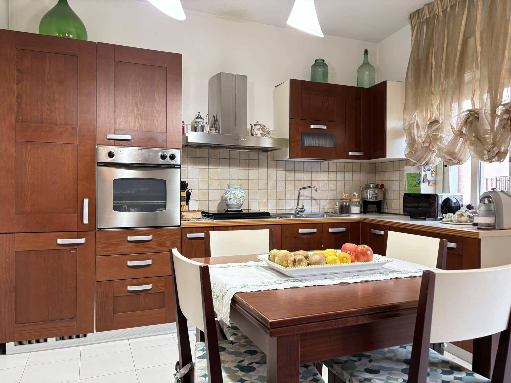 Pentolame, cucina moderna con mobili in legno, frigorifero, forno a microonde, tavolo da pranzo, sedie imbottite, dettagli decorativi, elettrodomestici, accessori per cucina, finestra con tende di pizzo, ambiente accogliente e funzionale per la casa.