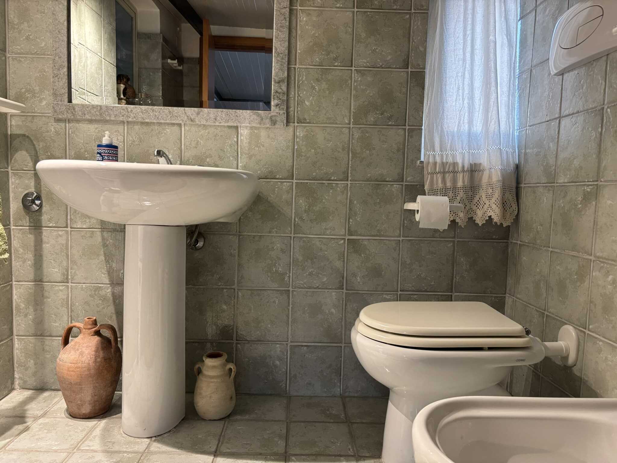 Lavandino e WC in un bagno moderno di casa con piastrelle di stile rustico, illuminate da luce naturale, con accessori eleganti e un aperto specchio sopra il lavandino, creando un ambiente accogliente e funzionale.