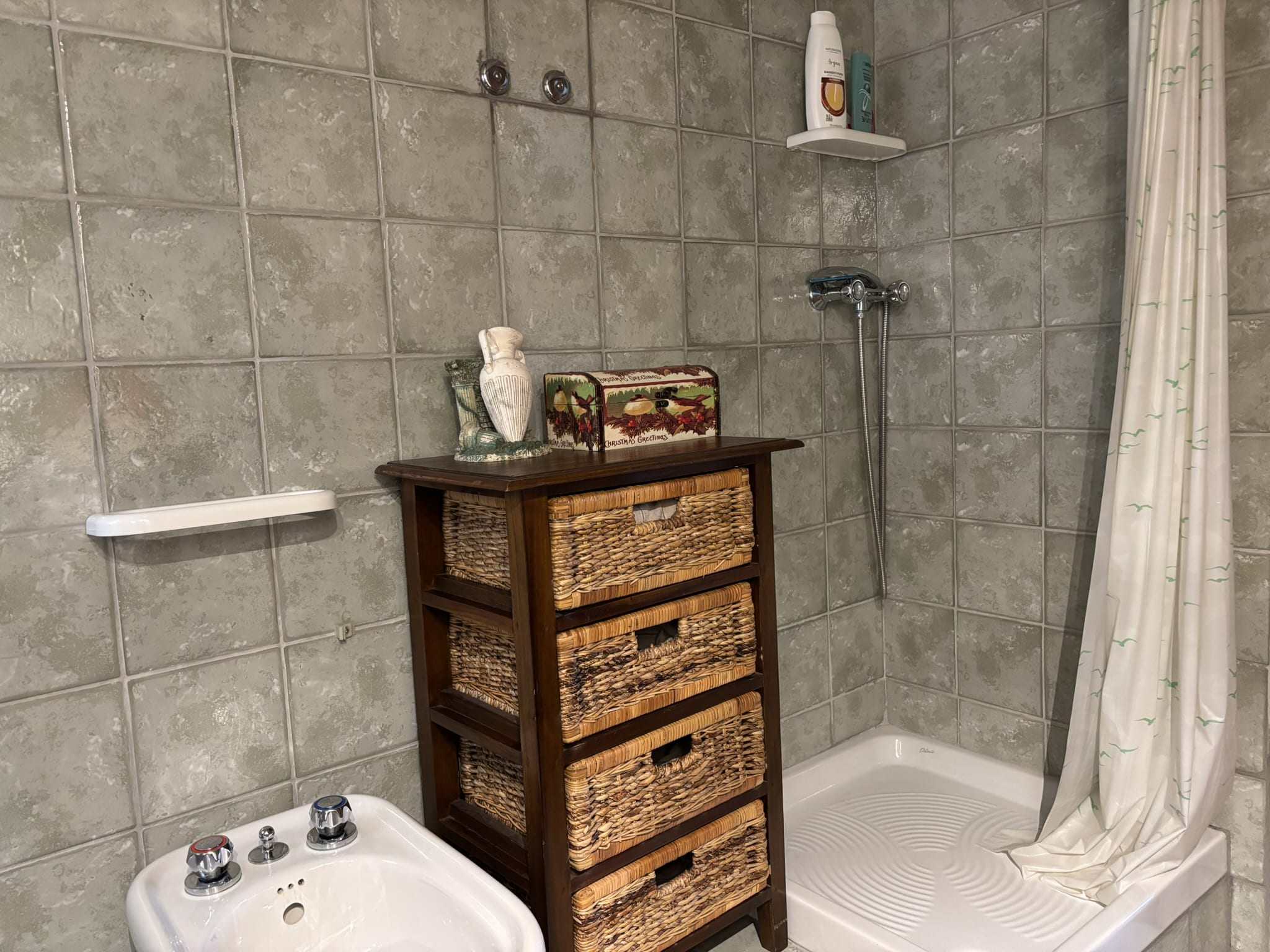 Lavabo e cabina doccia in un bagno con piastrelle grigie, armadietto in vimini, e accessori di cura personale, ideale per un ambiente accogliente e funzionale.