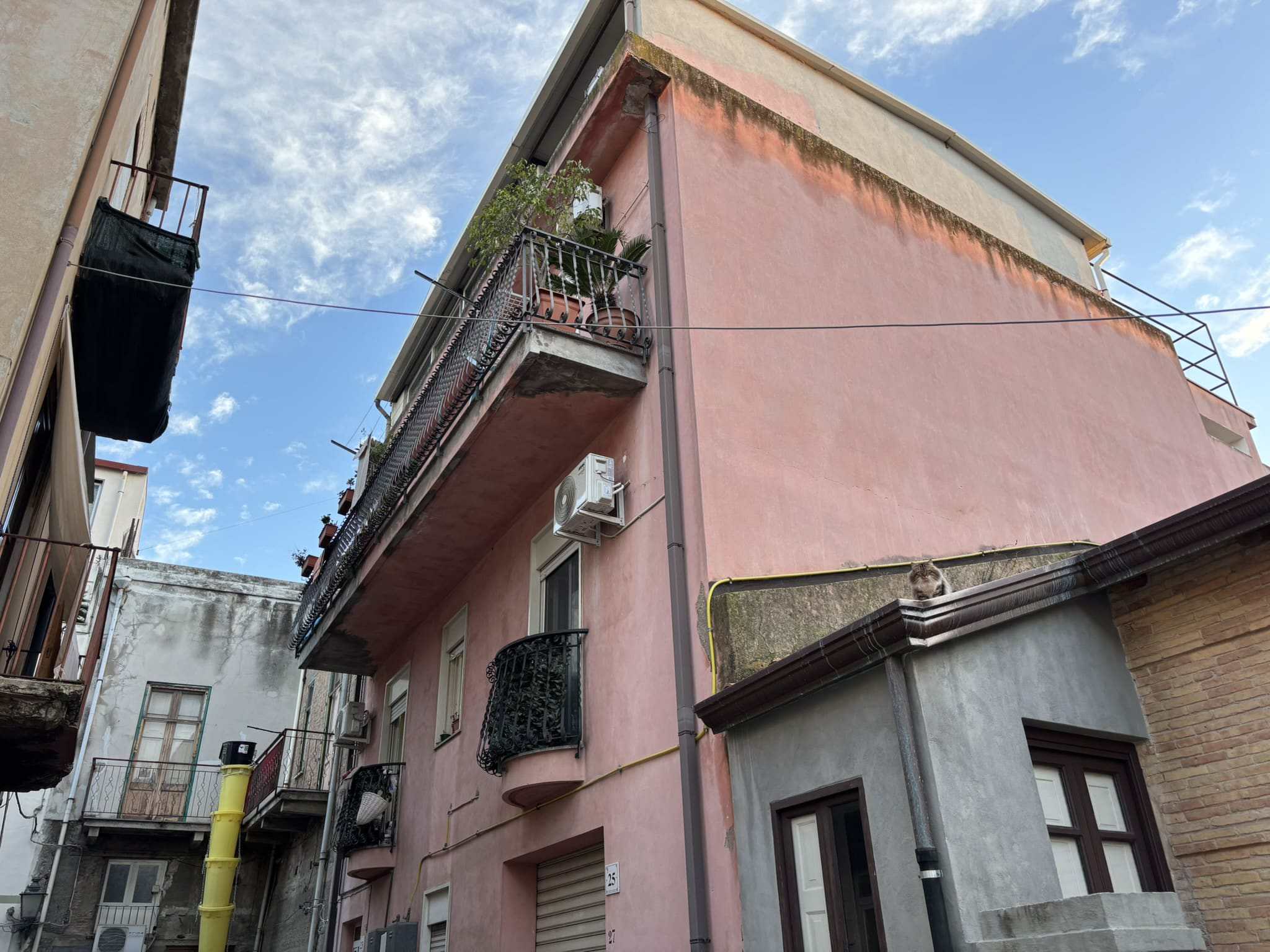 Balcone di un appartamento con piante, aria condizionata e ringhiera in ferro battuto, facciata esterna in colori pastello, in un contesto urbano, cielo azzurro con nuvole.