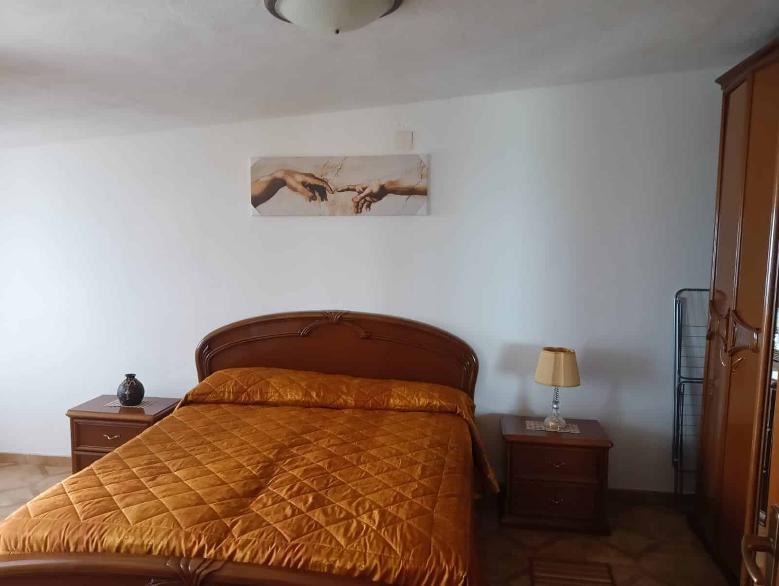 Letto matrimoniale in stile classico con copriletto color ocra, nelle dimensioni di una camera da letto spaziosa con mobili in legno, lampada da tavolo, armadio a due ante e decorazione artistica raffigurante le mani di Gesù.