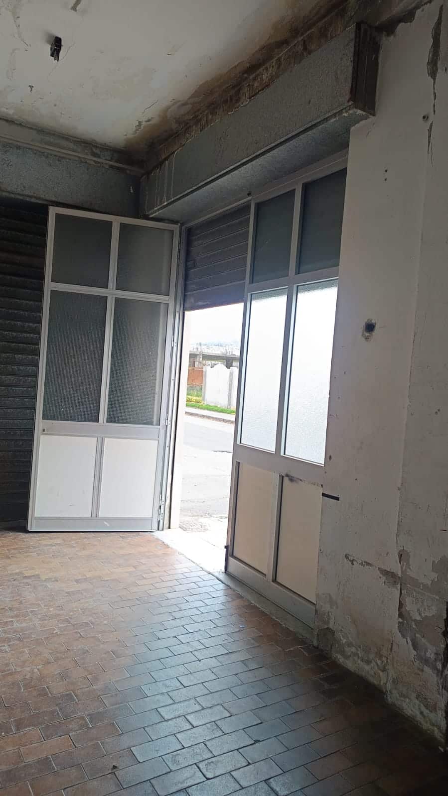 Porta d'ingresso in fibra di vetro con vetri trasparenti e persiana automatica, ambiente interno con pareti in muratura e pavimento in piastrelle in cotto, in fase di ristrutturazione.