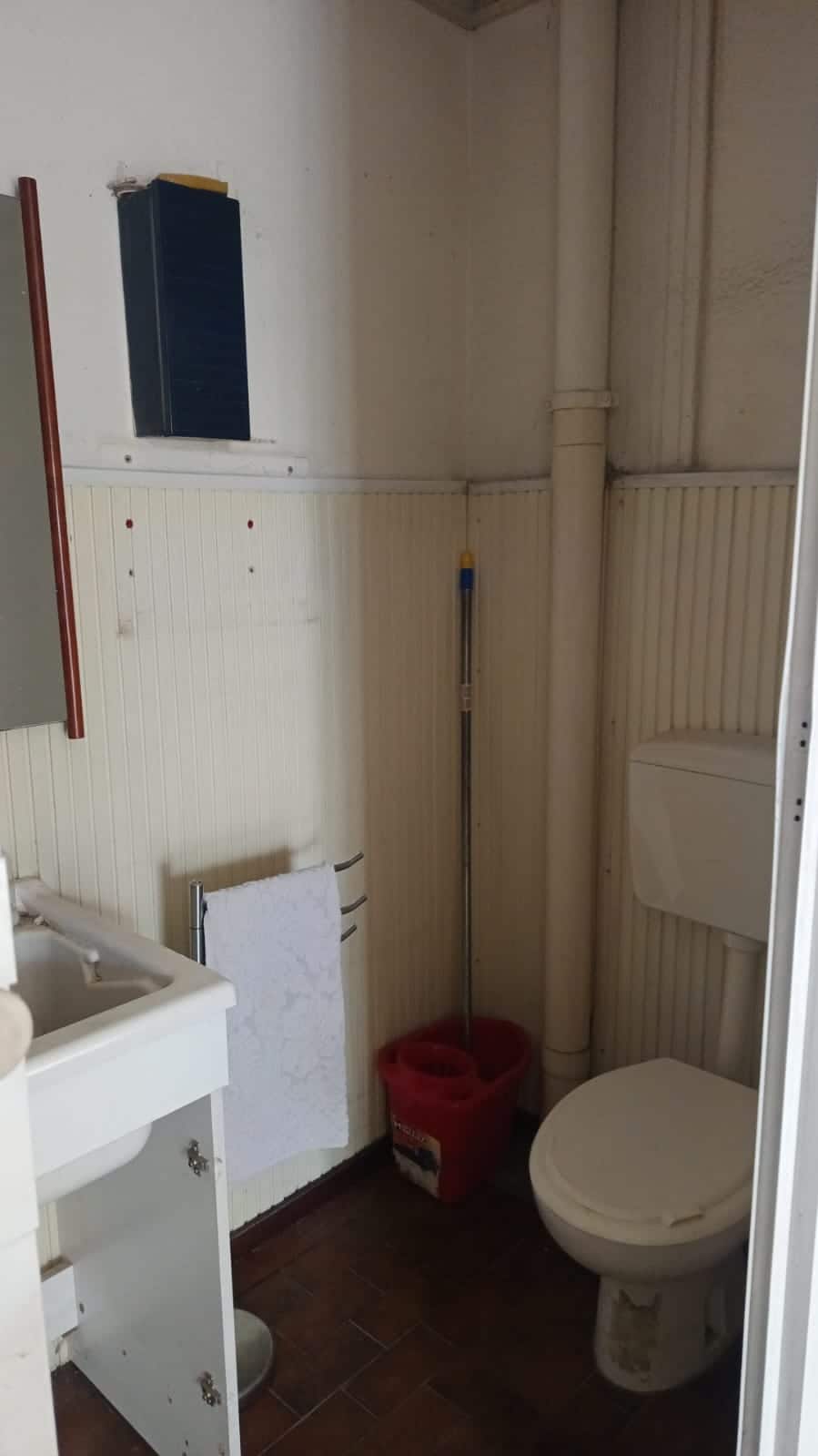 WC bagno piccolo con mobili essenziali, pannelli in plastica sulle pareti e dettagli pratici, come scopa e asciugamano, ideale per spazi ridotti o aggiunte rapide.