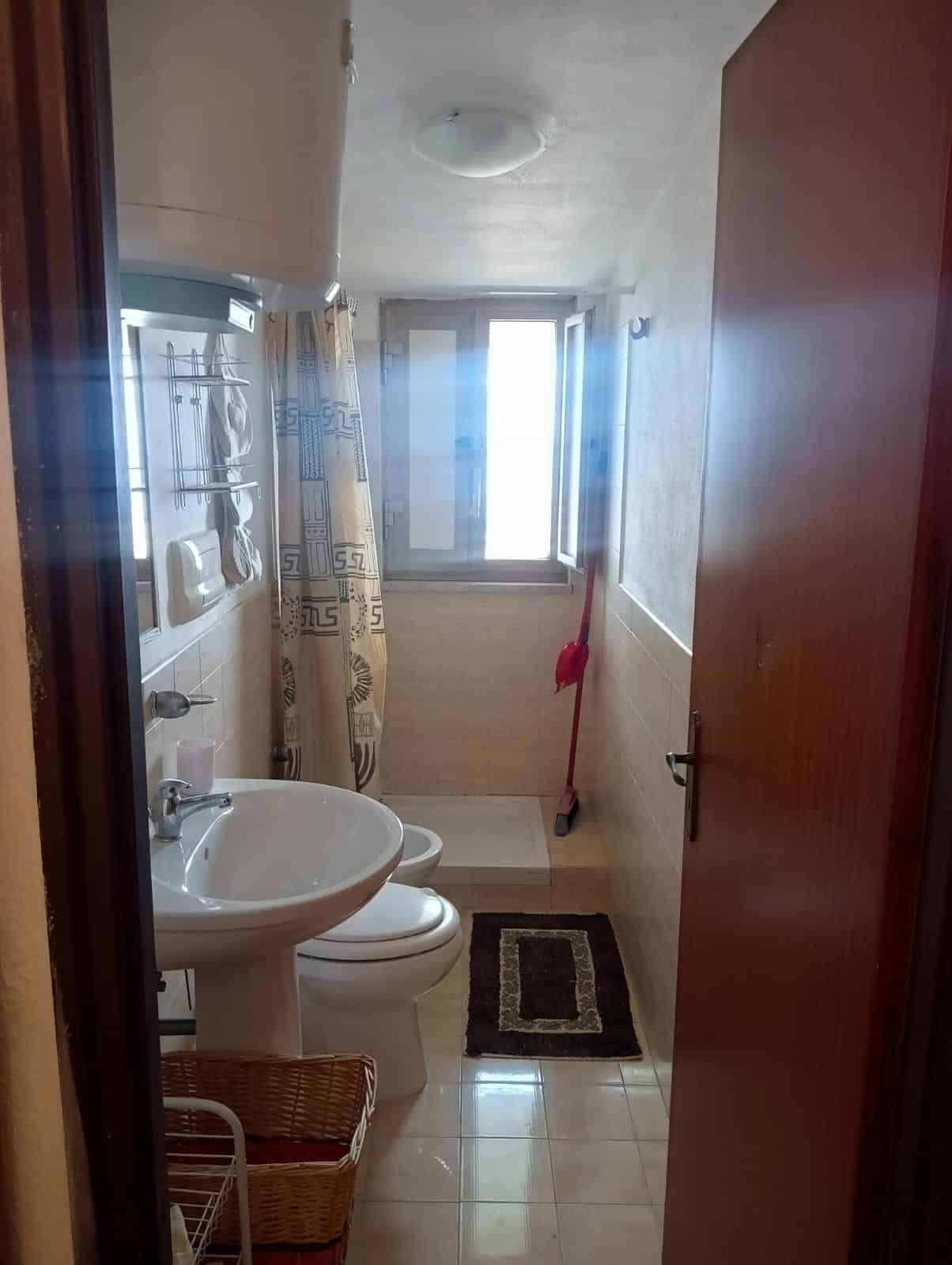 Lavabo e servizi igienici in bagno con finestra e tende svolazzanti, arredato con moquette e accessori come scopino e cesto porta oggetti, perfetto per servizi quotidiani.