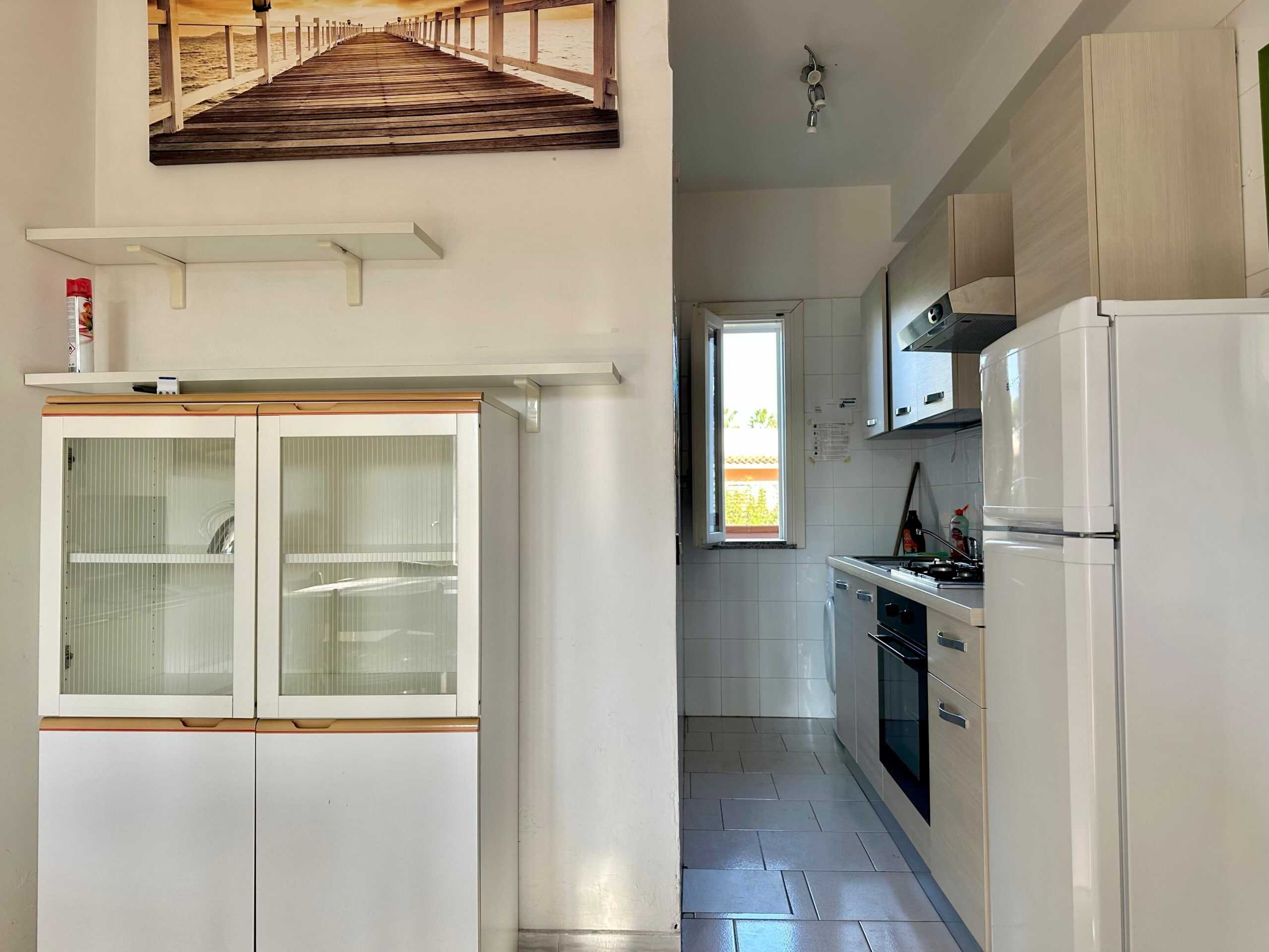 Armadio bianco con ante in vetro e mensole di scuola, cucina compatta con frigorifero, forno e piano cottura in stile moderno, finestra luminosa e pareti bianche.