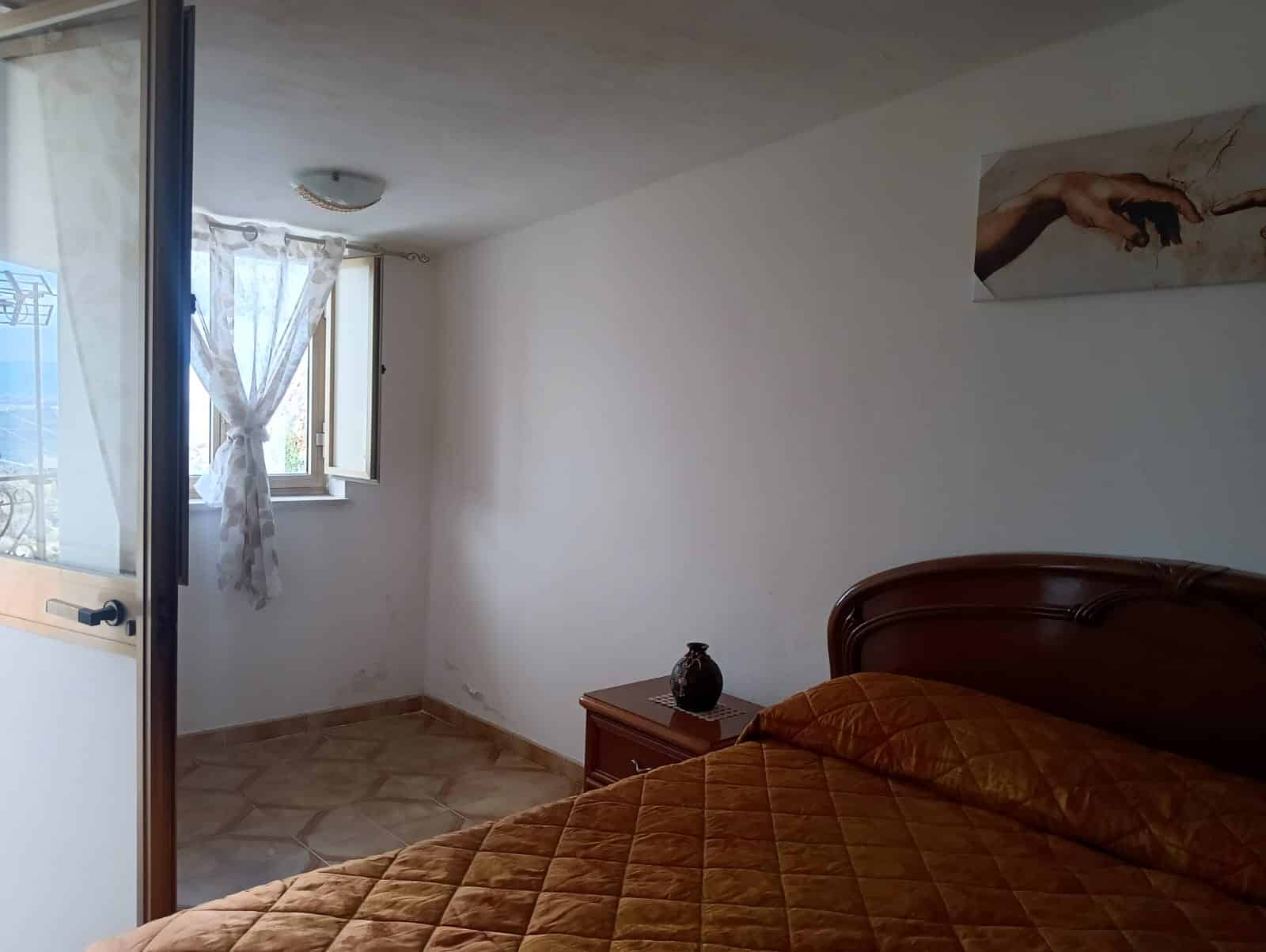 Camera da letto con pareti bianche, letto con rivestimento in velluto marrone, finestra con tende leggere, arredamento essenziale, ambiente luminoso e accogliente, ideale per appartamento in affitto o vendita.