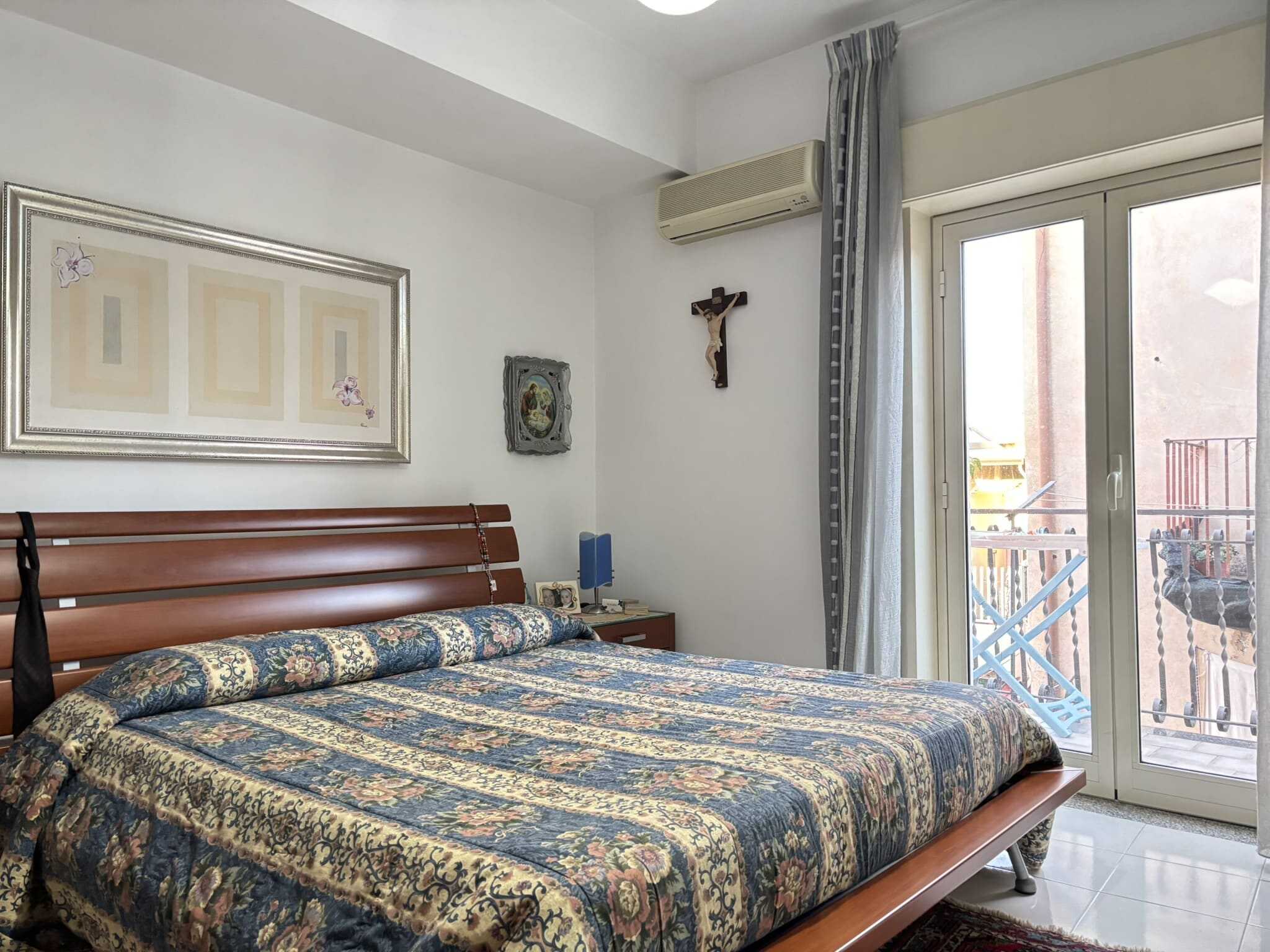 Letto matrimoniale in una camera da letto con arredi classici e tende a righe, balcone con vista urbana, ambiente luminoso e accogliente, stile semplice e funzionale in un appartamento in Italia.