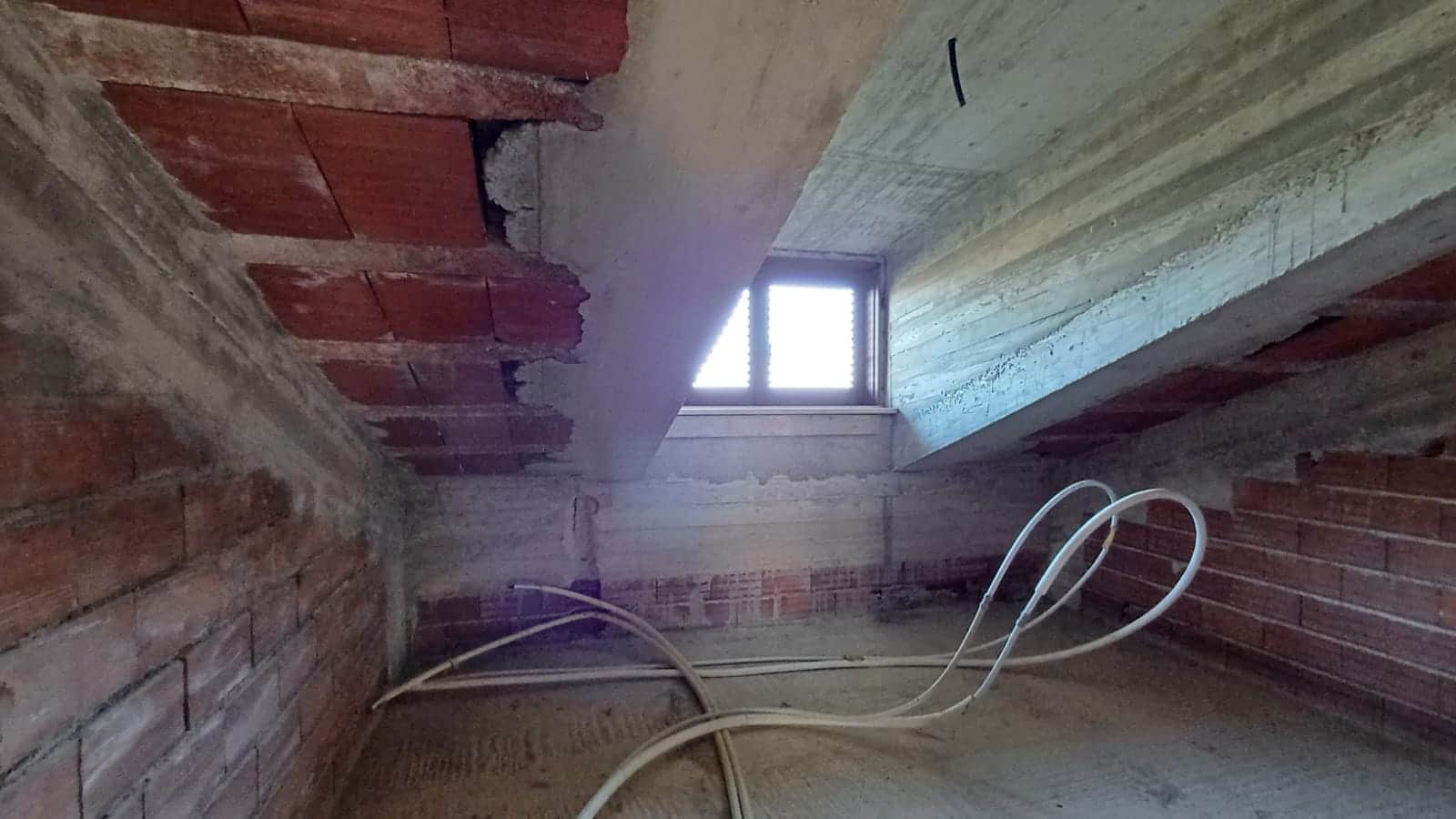 Finestra in mansarda in costruzione con pareti in mattoni e cemento, interno di casa in fase di ristrutturazione, prove di isolamento e impianto elettrico, atmosfera di lavoro edilizio.